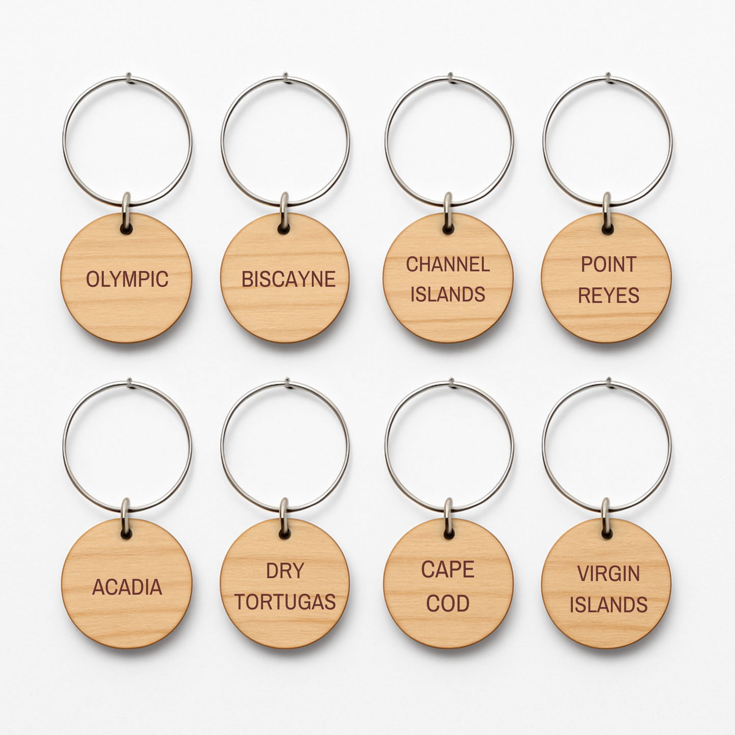 Eight round wooden wine charms with metal rings from Mirabilia Boutique’s Coastal Escapes Collection are arranged in two rows. Each charm is engraved with names of national parks like Olympic, Biscayne, and Channel Islands.