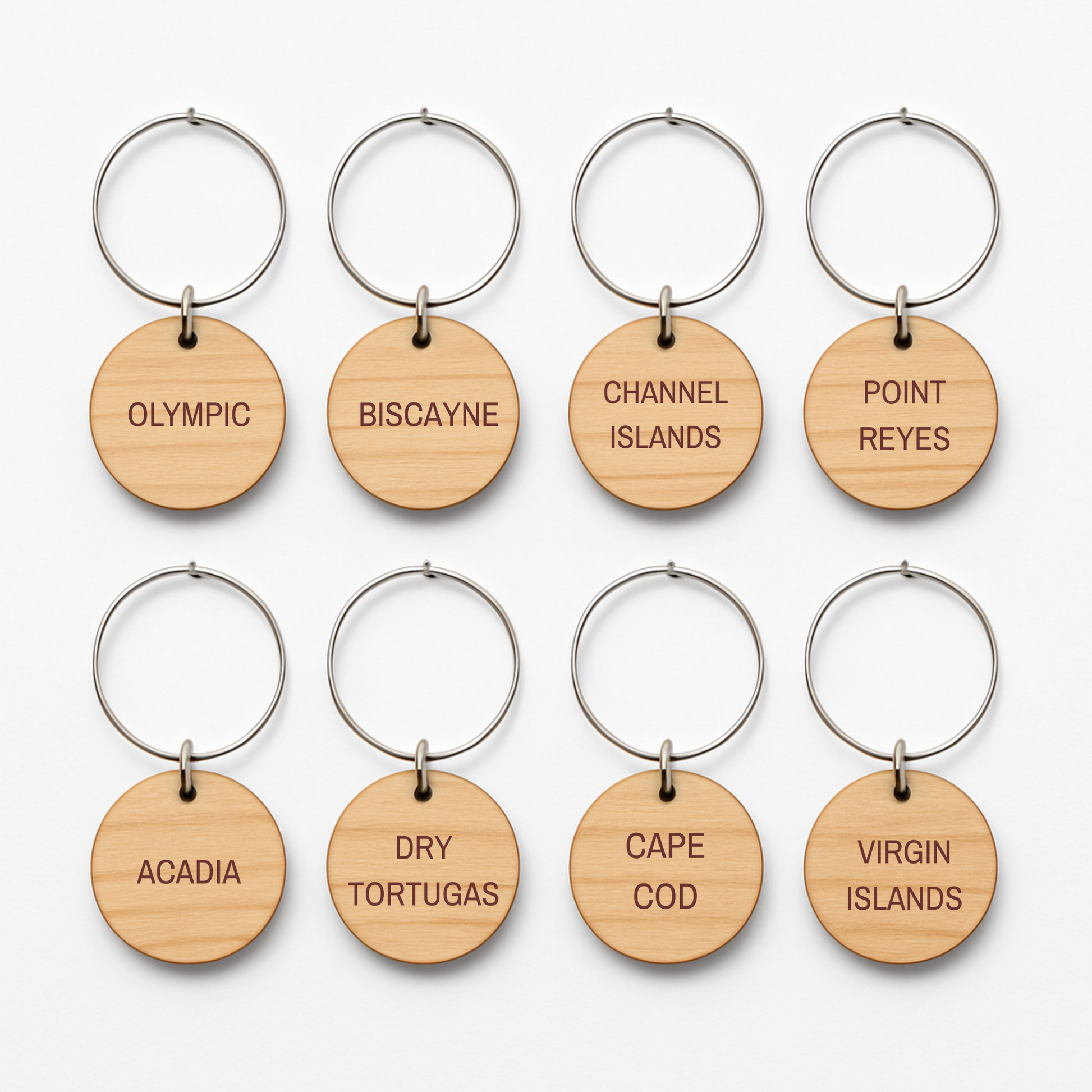 Eight round wooden wine charms with metal rings from Mirabilia Boutique’s Coastal Escapes Collection are arranged in two rows. Each charm is engraved with names of national parks like Olympic, Biscayne, and Channel Islands.