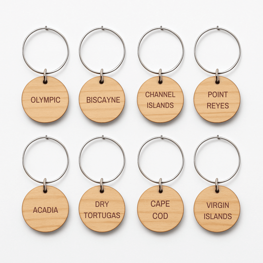 Eight round wooden wine charms with metal rings from Mirabilia Boutique’s Coastal Escapes Collection are arranged in two rows. Each charm is engraved with names of national parks like Olympic, Biscayne, and Channel Islands.
