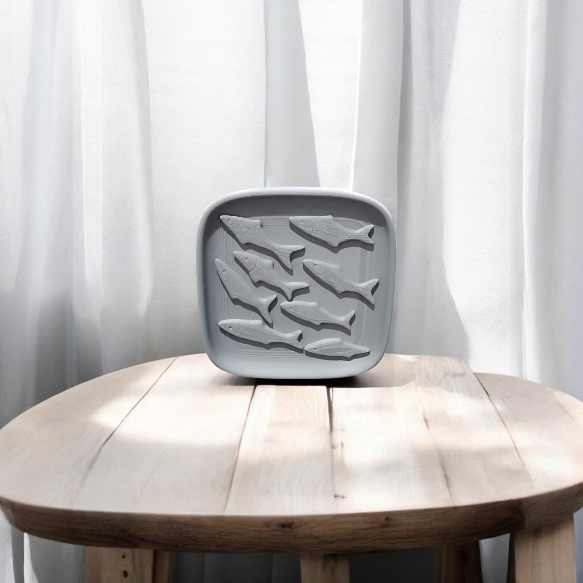The Coastal Fish Decorative Tray by Mirabilia Boutique, a gray silicone tray featuring six fish-shaped cavities, rests on a light wooden table before white curtains.