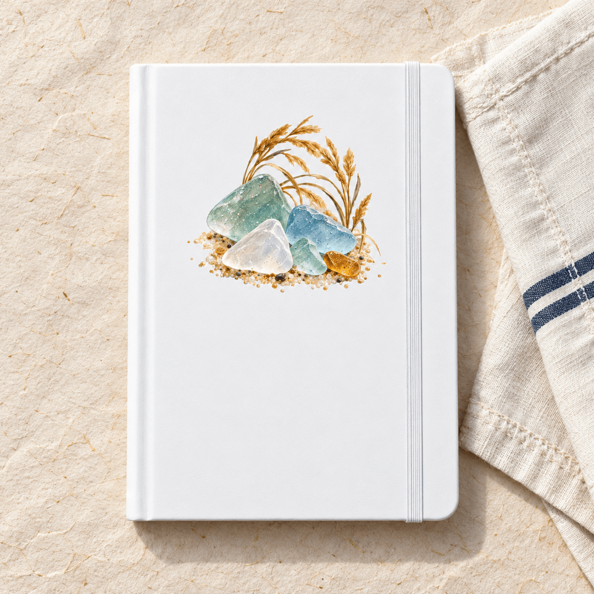 A white A5 A Little of the Shore Journal featuring a watercolor illustration of sea glass and wheat stalks on the cover, placed on a textured surface next to a cloth napkin.