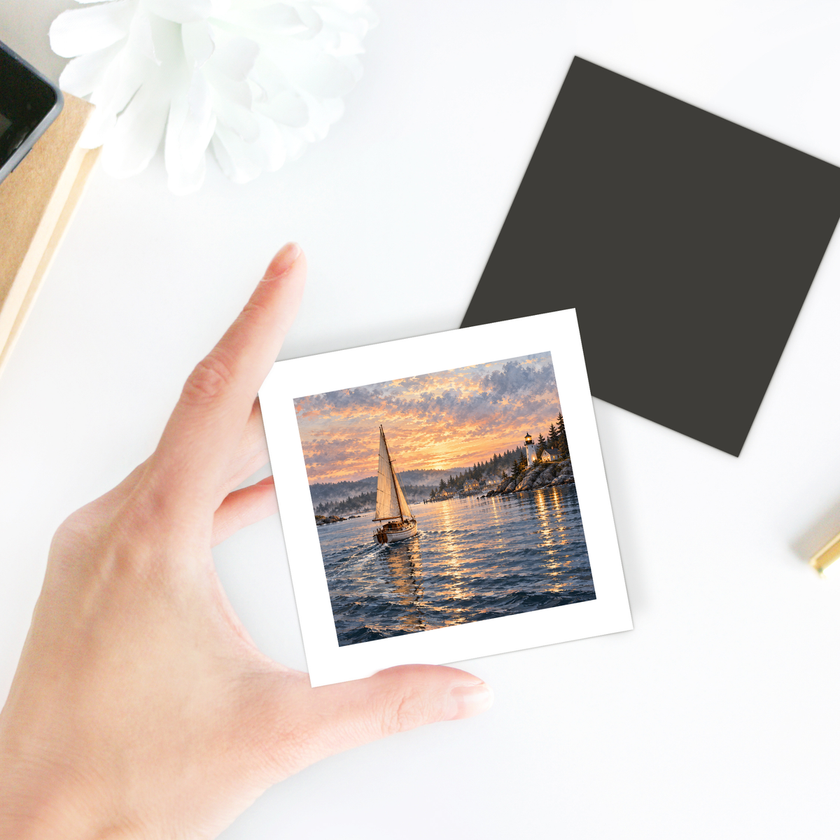 A hand holds The Long Way Home Magnet, a square decorative hardboard magnet featuring a sailboat on the water near a lighthouse at sunset.