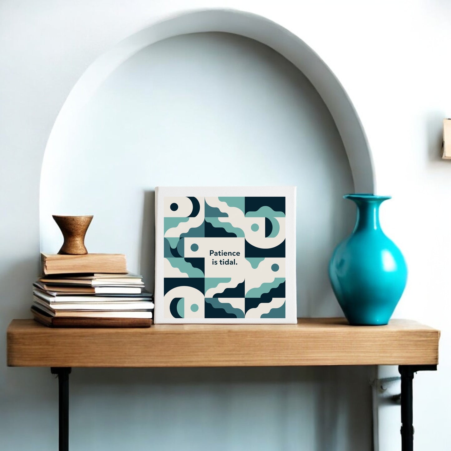A wooden shelf with stacked books, a small cup, a turquoise vase, and Mirabilia Boutique’s "Patience Is Tidal Canvas – Calm Coastal Wall Art" sits before a white arched alcove.
