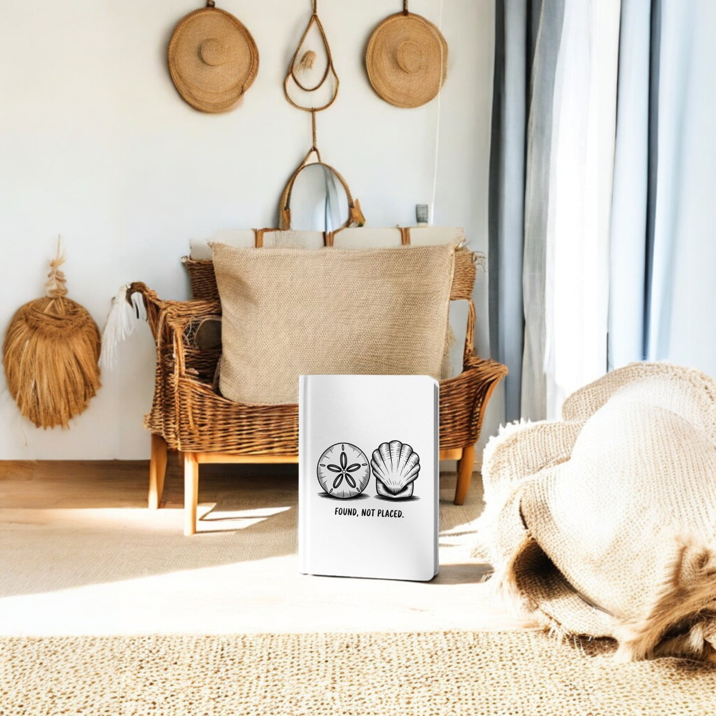 A wicker chair with beige pillows sits in a sunlit room. The Mirabilia Boutique "Found, Not Placed" Coastal Quote Notebook stands on a light rug, adding artistic charm and coastal style to the serene space.