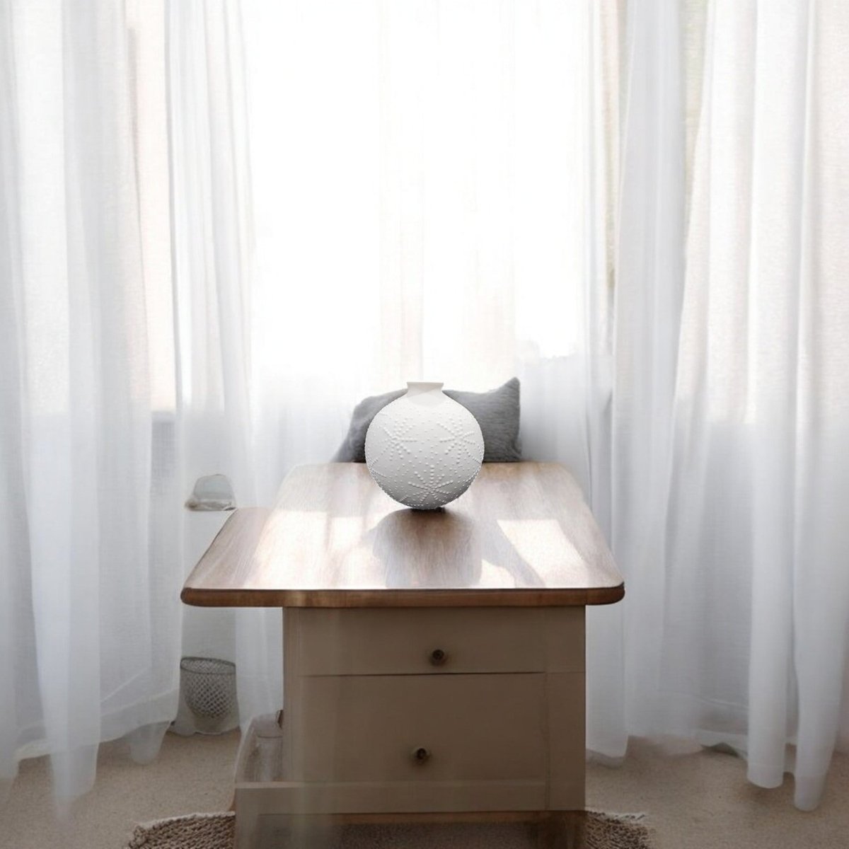 The Sand Dollar Textured Vase in Matte White by Mirabilia Boutique sits on a wooden table with a beige drawer, in front of sheer white curtains, its coastal-inspired design enhancing the calm, minimalist ambiance with soft natural light.