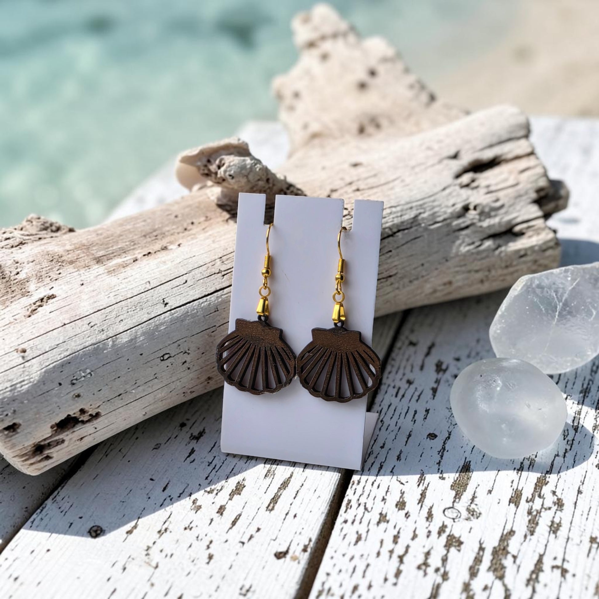 Scallop Shell Earrings | Lasercut Walnut Wood by Mirabilia Boutique—gold-hook earrings displayed on a white card over rustic wood, driftwood, and crystals, with a blurred beach view. Perfect coastal jewelry for ocean lovers.