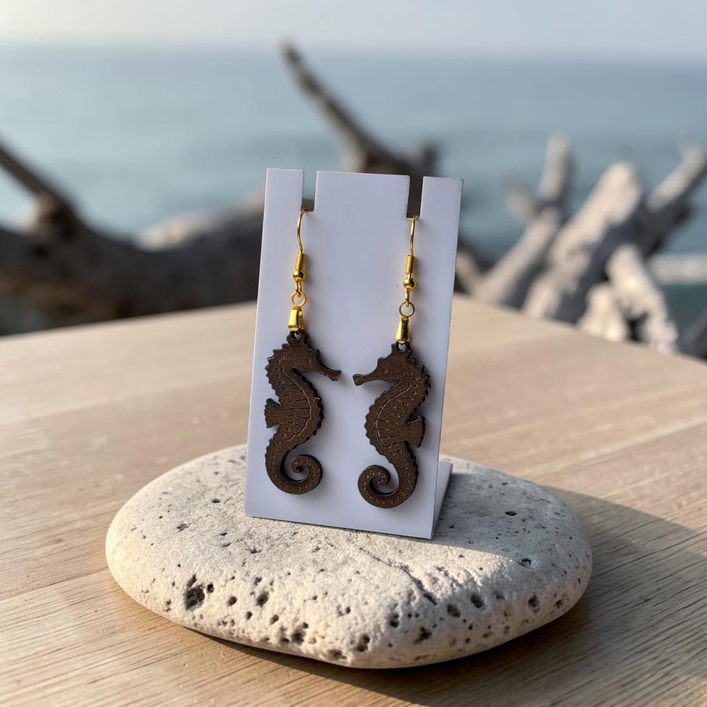 Seahorse Earrings | Lasercut Walnut Wood by Mirabilia Boutique rest on a white card atop stone, with ocean, driftwood, and sky in the background—capturing the charm of unique ocean jewelry with elegant gold hooks.
