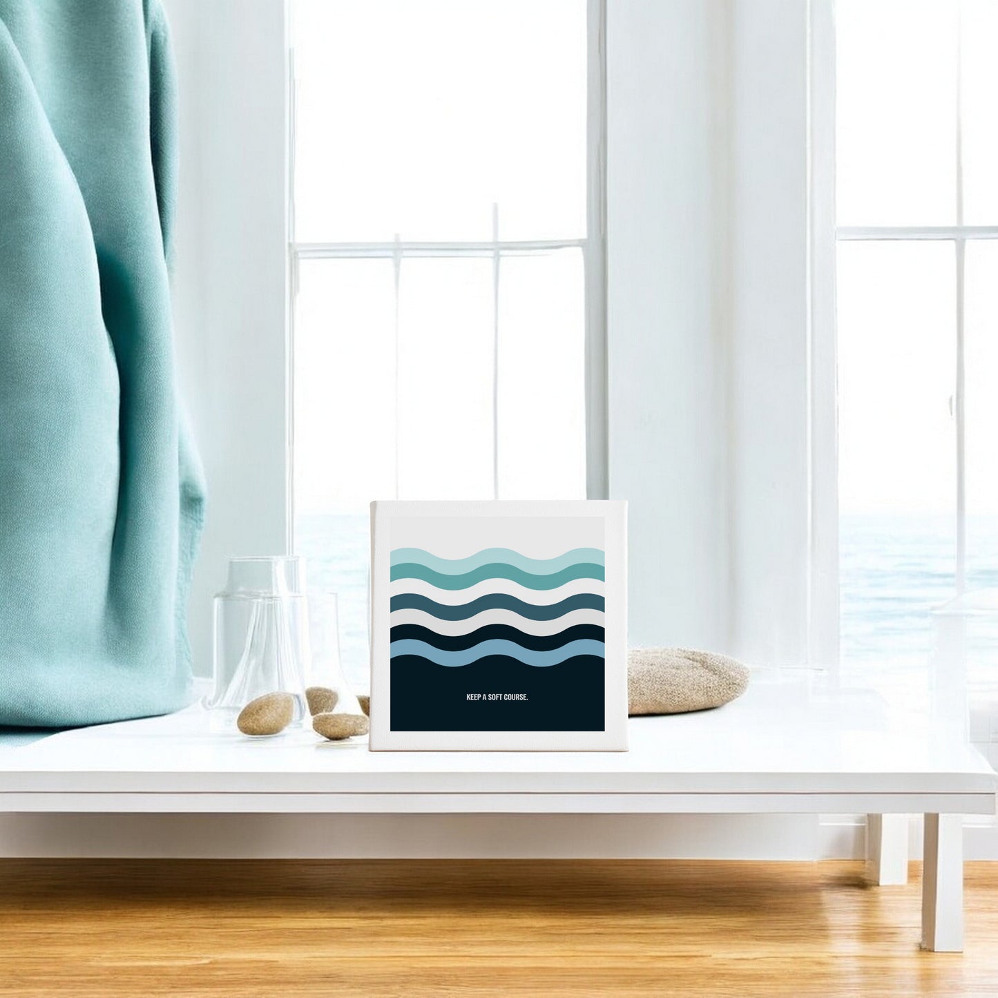 A minimalistic room features a white coffee table with calming decor—smooth stones and a folded blue blanket—while Mirabilia Boutique's "Keep a Soft Course Canvas Sign – 8x8 Minimal Ocean Art" hangs on the wall, and large windows reveal an airy ocean view.
