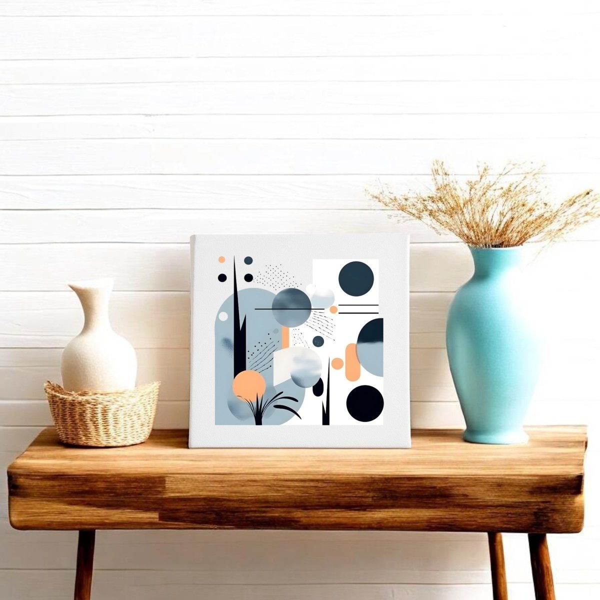 A wooden table displays a white vase in a wicker holder, a blue vase with dried grass, and the Coastal Sunrise Mid Century Modern Canvas by Mirabilia Boutique—an abstract sunrise art piece with geometric shapes—set against a white shiplap wall.