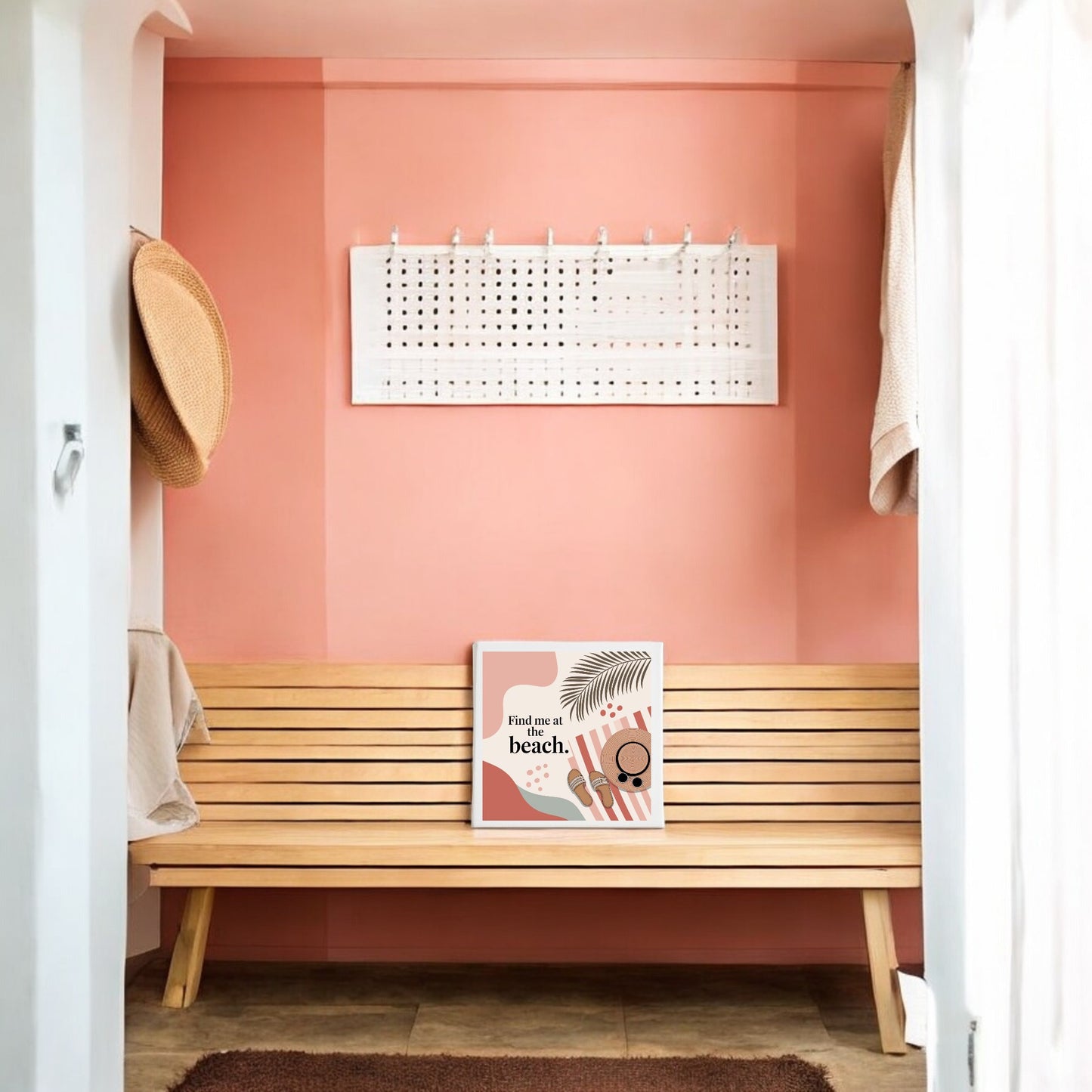 A wooden bench sits against a pink hallway wall. Above is a white pegboard with hooks. The bench displays the Mirabilia Boutique “Find Me at the Beach Canvas – Coastal Summer Vibes,” while a woven hat hangs on the left wall.