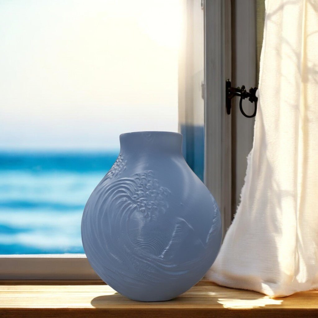 The Stone Blue Coastal Wave Vase by Mirabilia Boutique rests on a wooden sill. Sunlight highlights its subtle wave patterns, framing a serene ocean view—ideal for coastal decor. A white curtain hangs softly to the right.