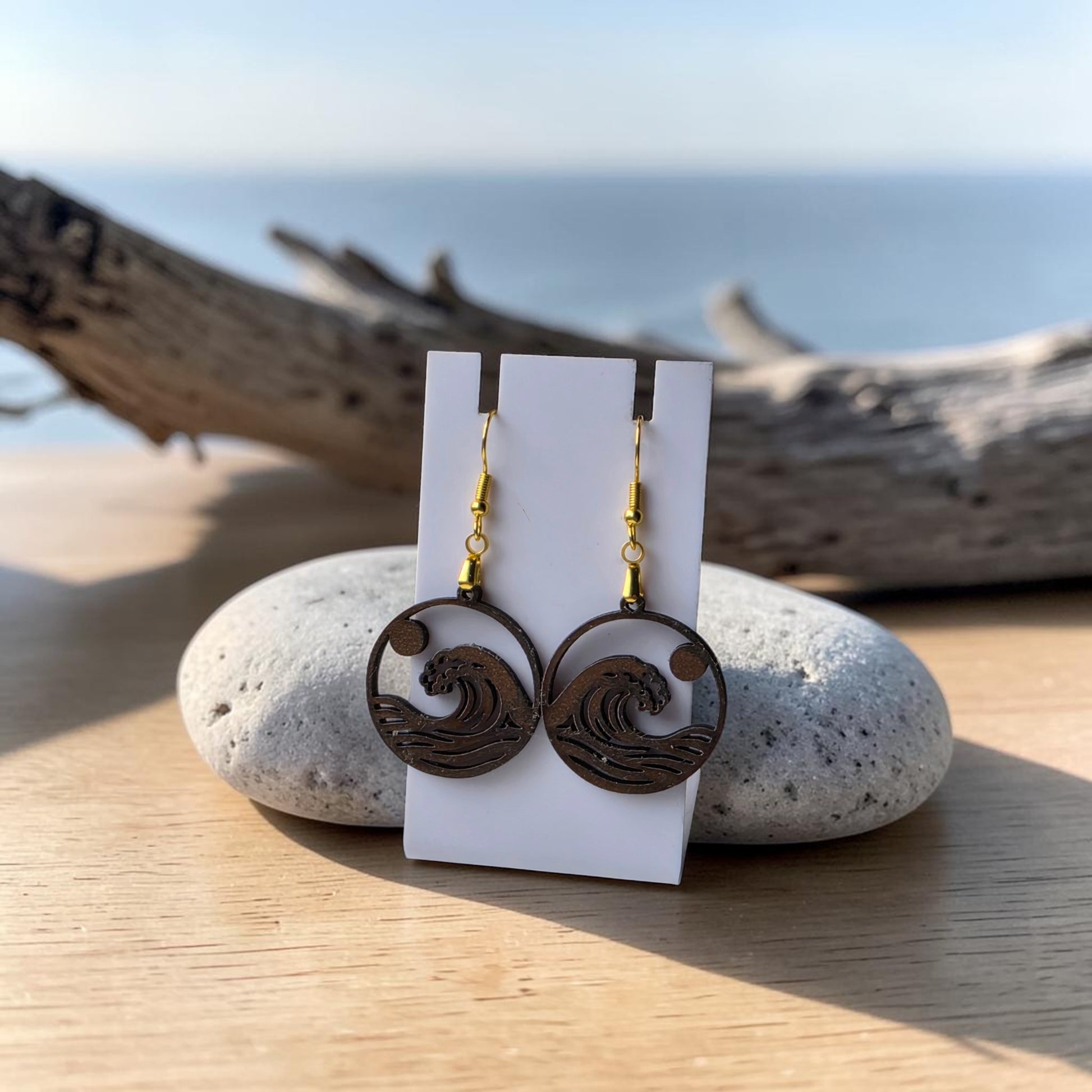 The Sunrise Wave Earrings by Mirabilia Boutique are lasercut from walnut wood, shown on a white card atop a gray stone with driftwood and the sea behind—perfect coastal jewelry.