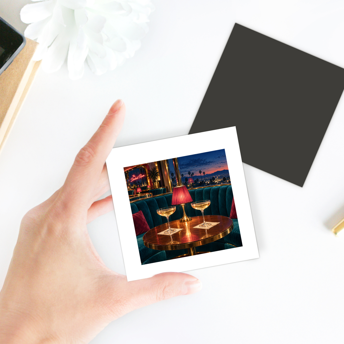A hand holds a square Cocktails at the Stardust Magnet | Chrome & Starlight featuring a photo of two drinks on a table inside a dimly lit lounge with a neon sign and a city sunset view.