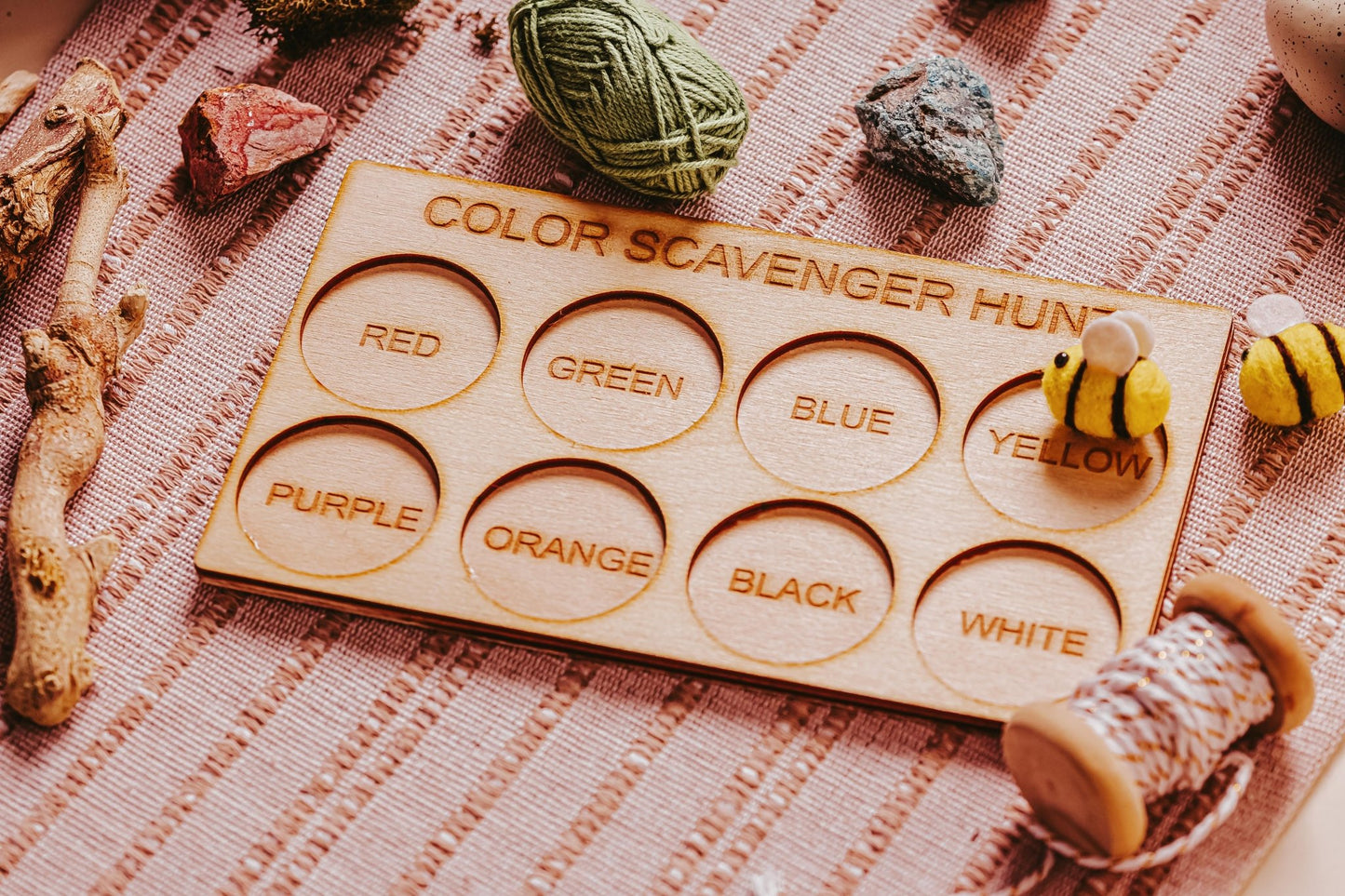 The "Exciting Color Scavenger Hunt Game for Kids' Outdoor Fun" by Mirabilia Boutique is an eco-friendly wooden board with circular cutouts in vibrant colors such as red, green, and blue. It comes surrounded by craft materials like yarn, rocks, wooden beads, and toy bees on a woven cloth. This outdoor toy is perfect for nature-inspired fun!