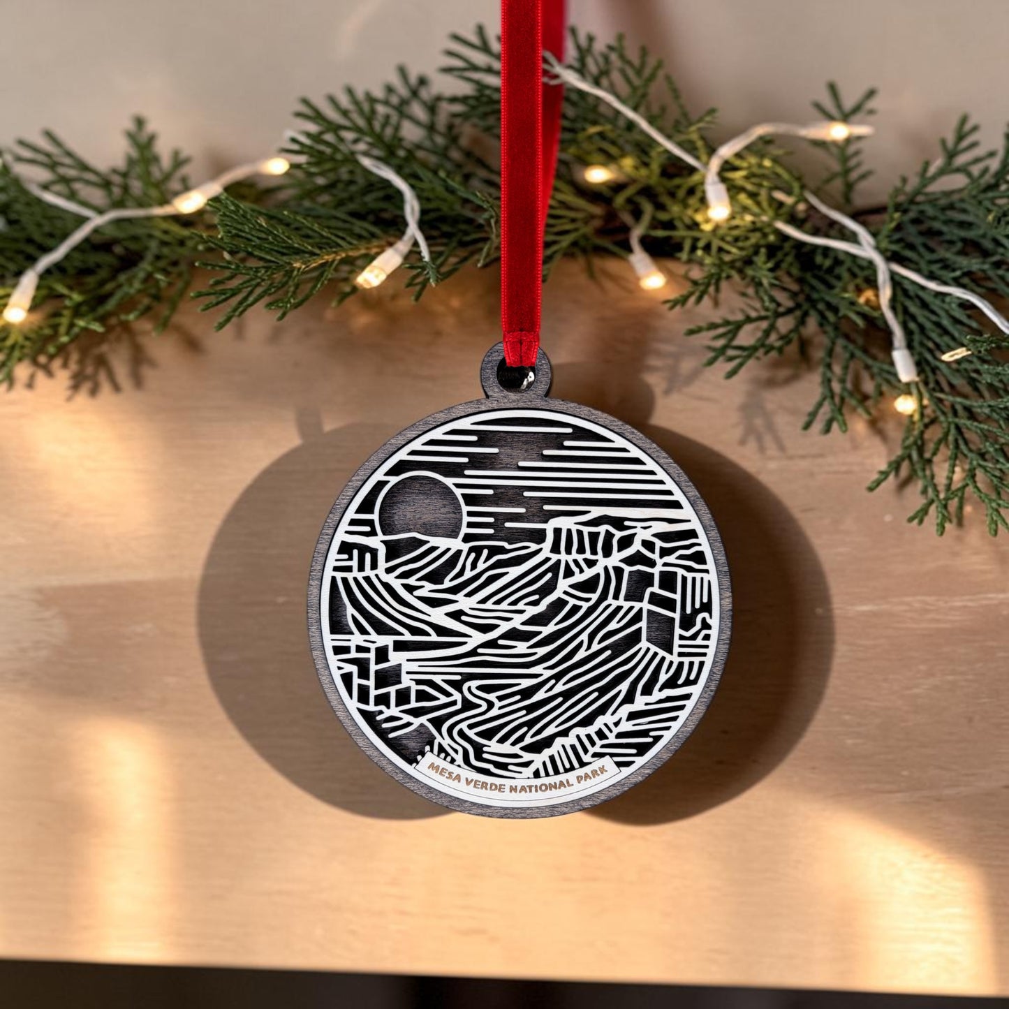 The Mesa Verde National Park Line Art Christmas Ornament by Mirabilia Boutique is a handcrafted wooden keepsake featuring a bold black and white maze design. It hangs from a red ribbon against pine branches and string lights, evoking the ancient cliff dwellings of the park.