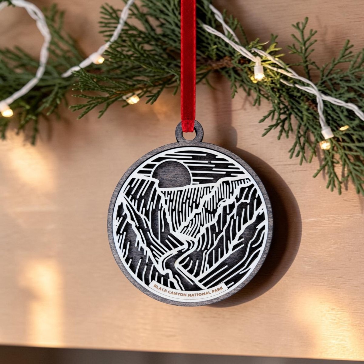 The Black Canyon of The Gunnison National Park Christmas Ornament by Mirabilia Boutique is a round, handcrafted wooden decoration featuring a striking black and white line art illustration of the park’s mountains and sun. It hangs from a festive red ribbon, while evergreen branches adorned with softly twinkling string lights create a cozy holiday backdrop.