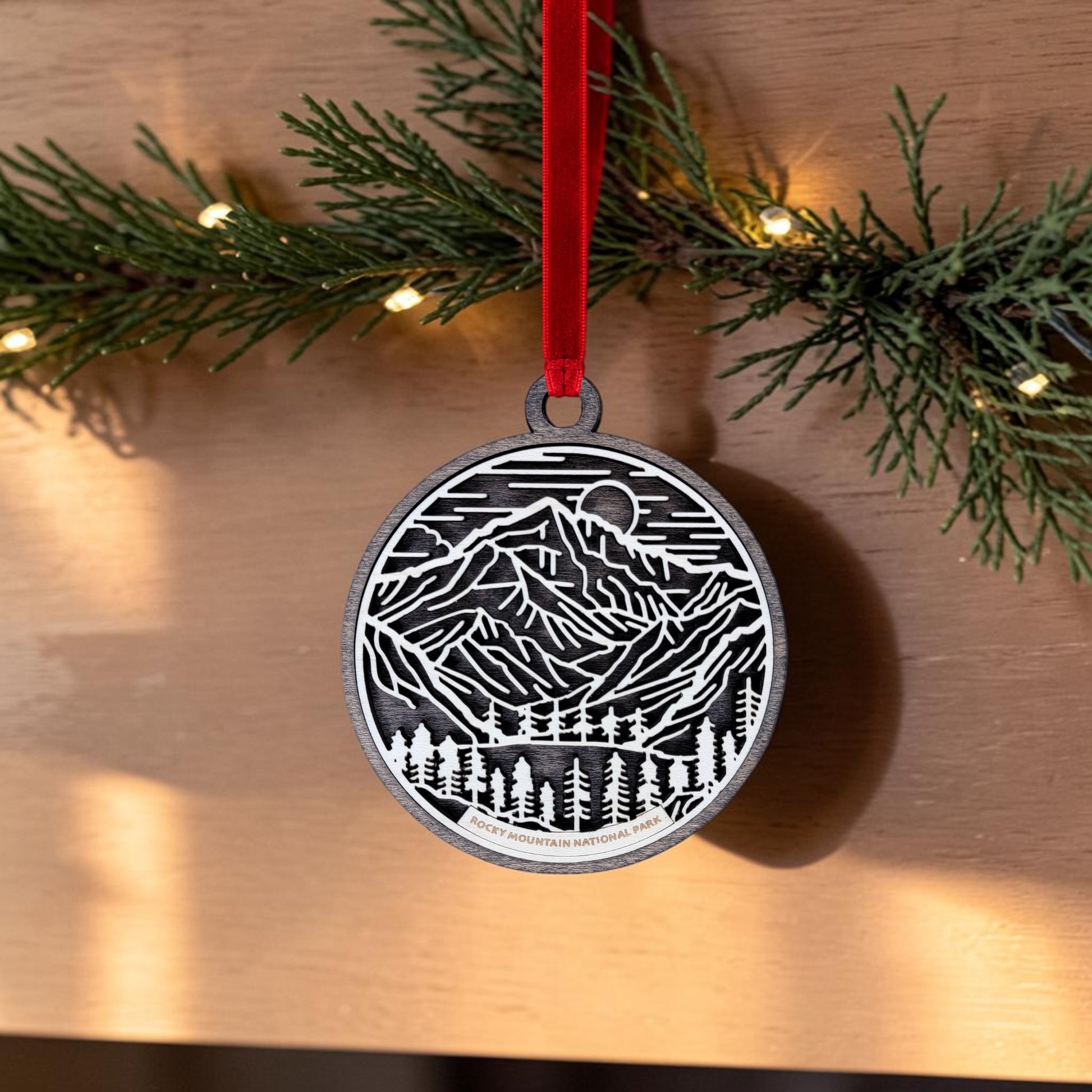 The Rocky Mountain National Park Line Art Christmas Ornament by Mirabilia Boutique, an oval wooden ornament with a black and white mountain and forest scene, hangs from a red ribbon on a branch adorned with small white lights in the background.