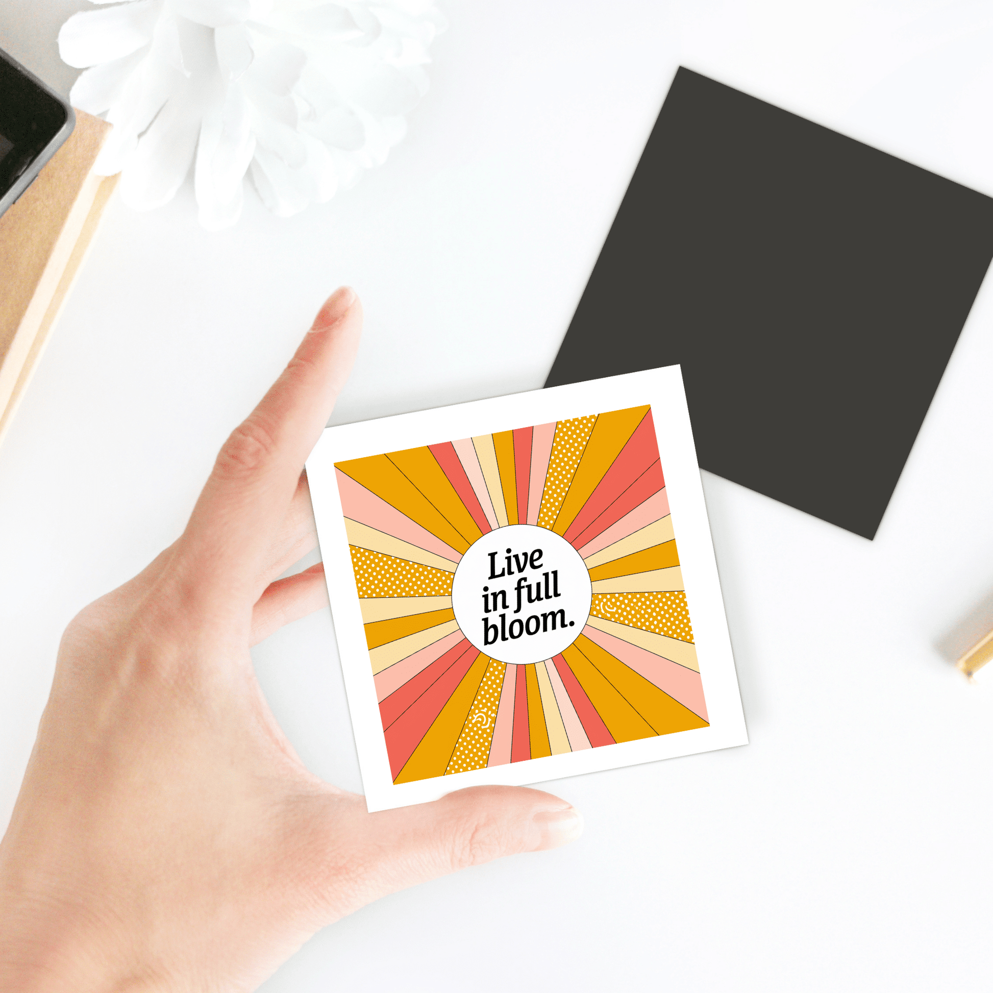 A hand holds the Mirabilia Boutique "Live in Full Bloom Magnet – Summer Sunburst Mini Art," featuring vibrant orange, yellow, and pink rays radiating from a circle with uplifting text—a perfect retro fridge magnet gift.