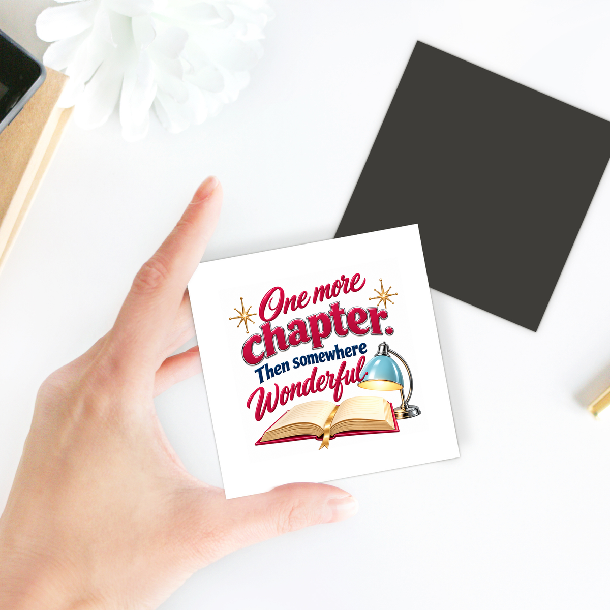 A hand holds a One More Chapter Book Lover Magnet | Chrome & Starlight featuring an open book, a desk lamp, and the text “One more Chapter. Then somewhere Wonderful.”.