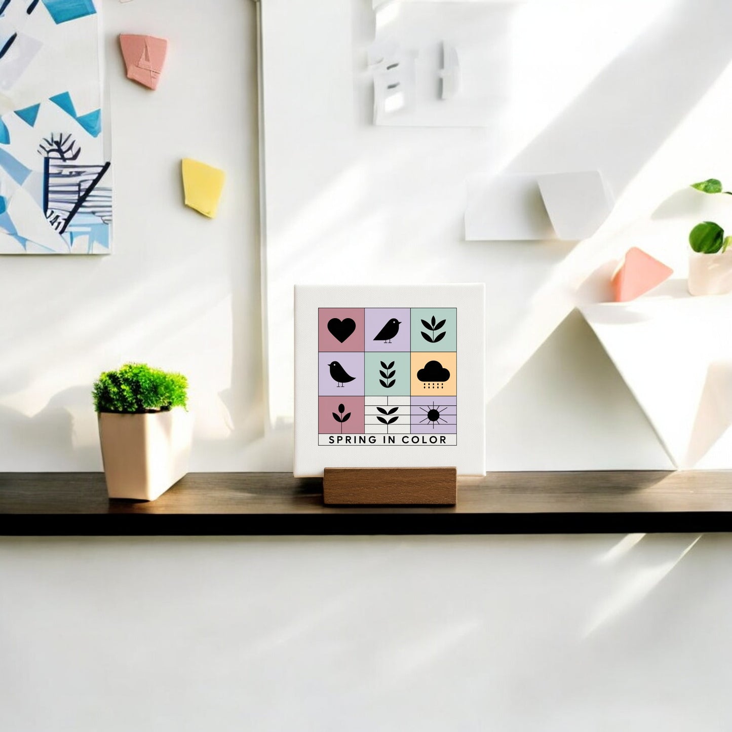 The Mirabilia Boutique "Spring in Color" Mini Canvas Sign with Wood Base adds a cheerful touch to a wooden shelf, especially when paired with a potted plant and geometric wall art for bright, modern spring décor.