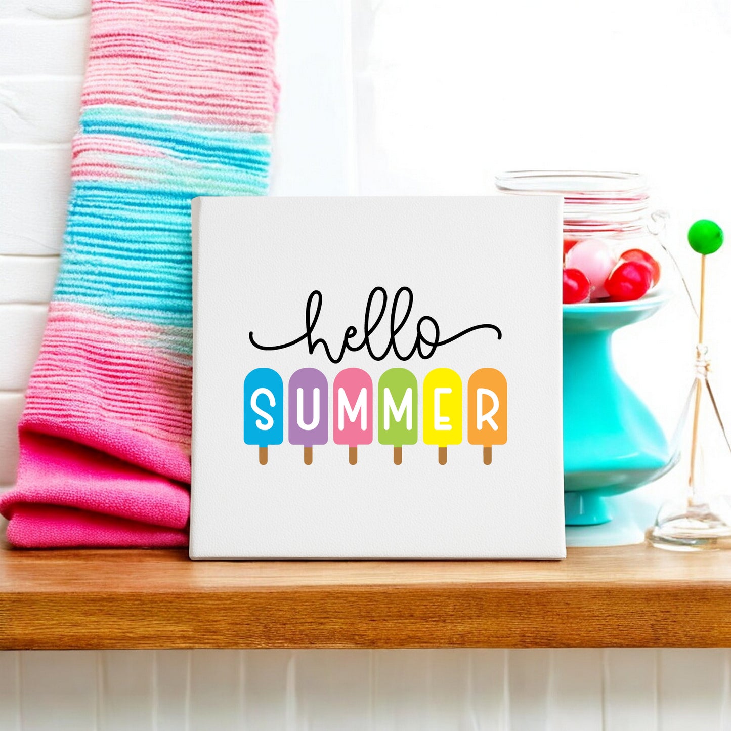 The Mirabilia Boutique Hello Summer Popsicle Canvas Sign (8x8), featuring “summer” on colorful popsicles, sits on a shelf with a pink and blue striped towel and a jar of red candies—cheerful decor for the season.