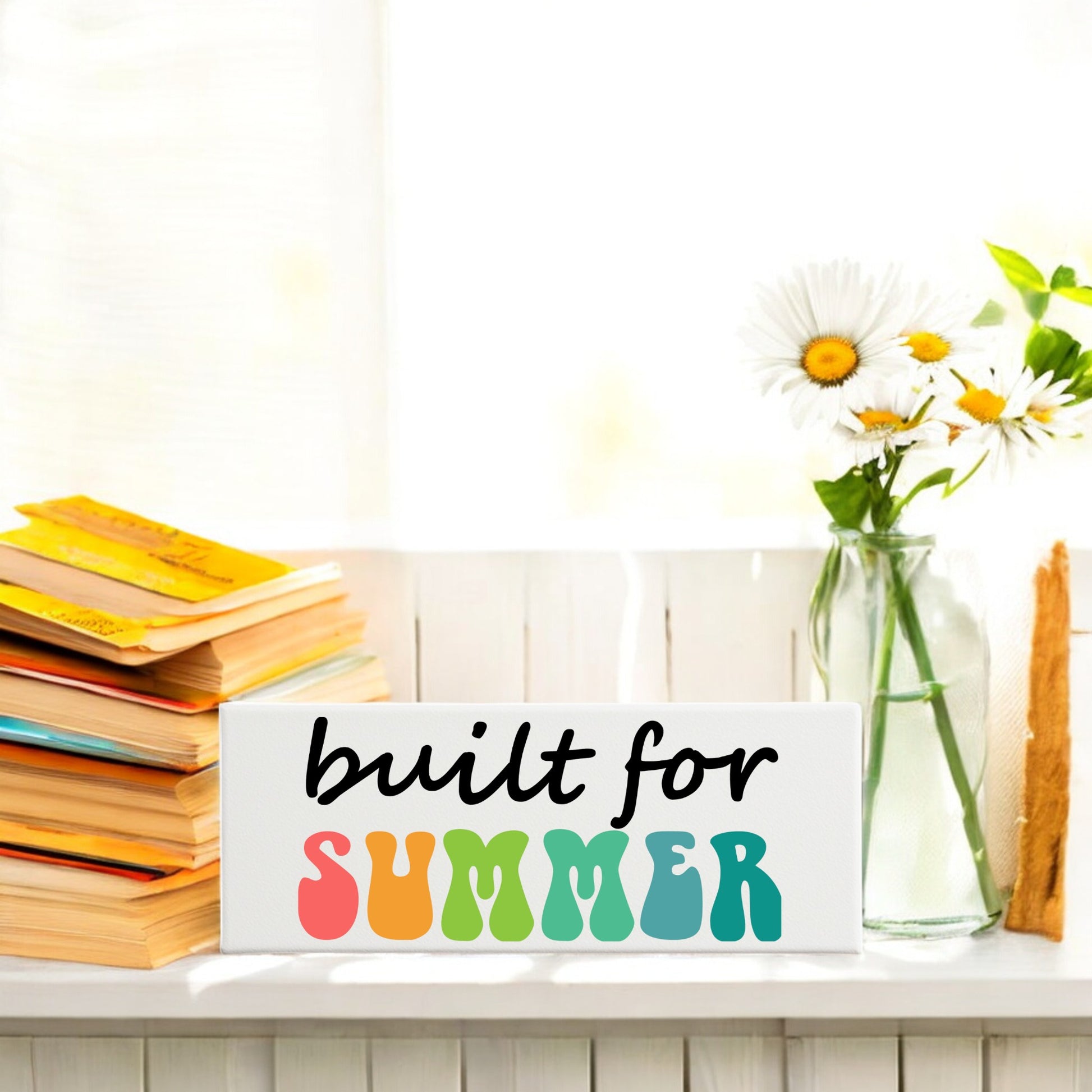 A stack of books, a glass vase with daisies, and the Mirabilia Boutique "Built for Summer Canvas – Retro Shelf Sign" are displayed on a white windowsill in bright sunlight.