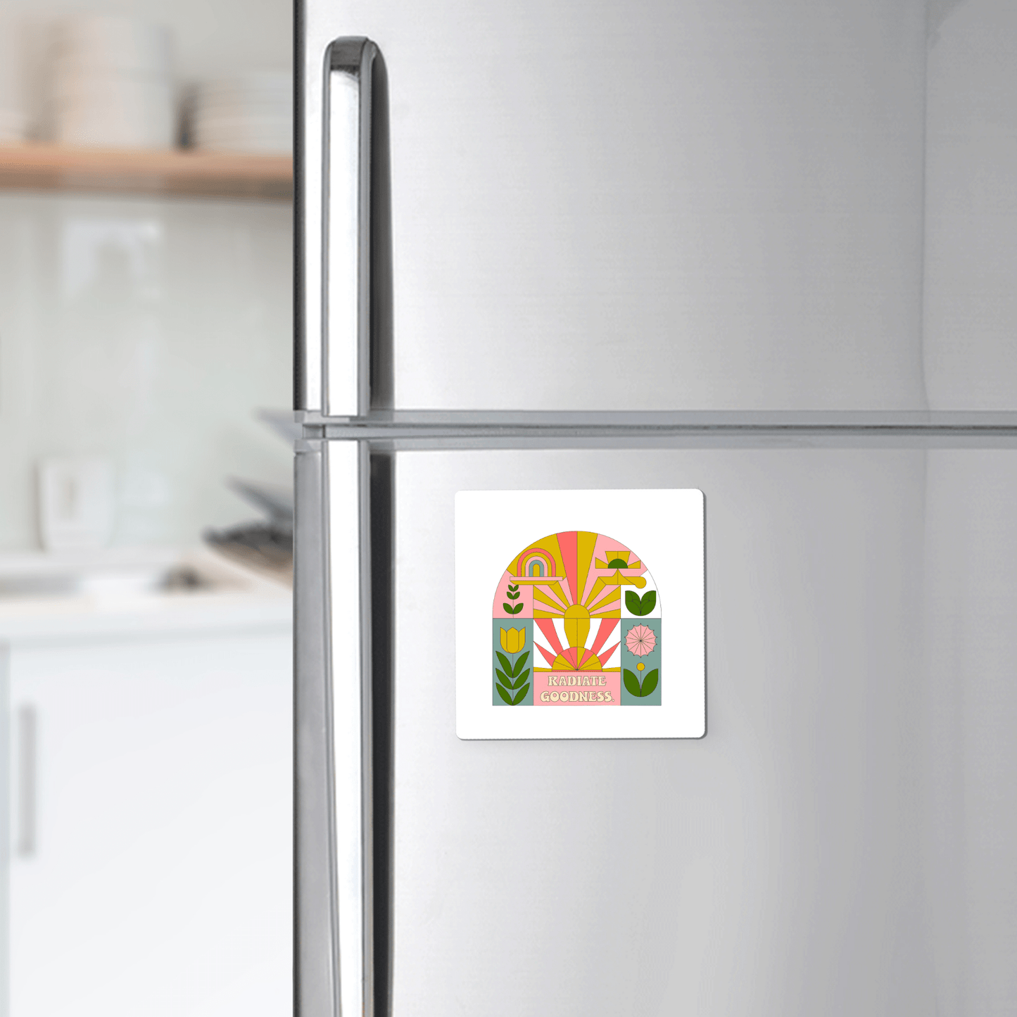 A silver refrigerator features Mirabilia Boutique's Radiate Goodness Colorful Floral Magnet showcasing abstract shapes, leaves, flowers, and the words "RADIATE GOODNESS," set against a bright, tidy kitchen backdrop.