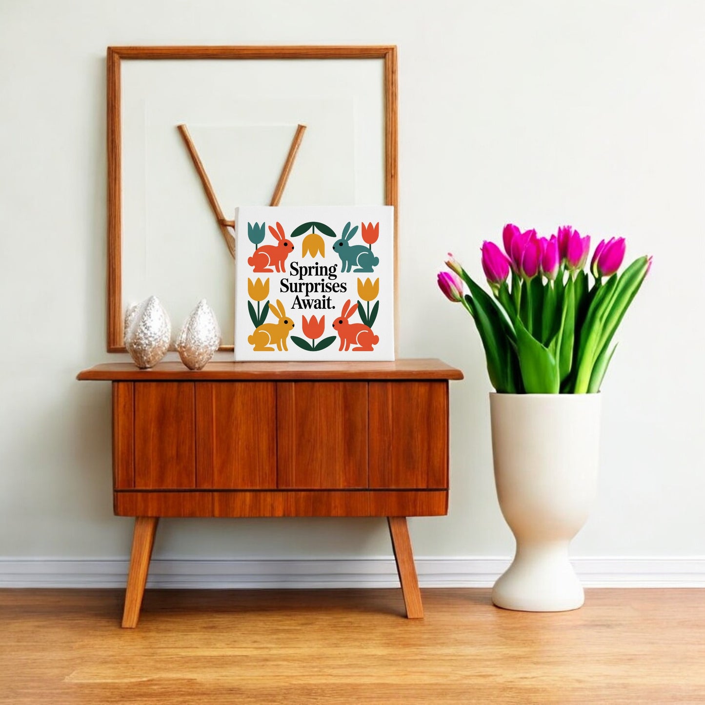 A wooden sideboard with decor sits by a wall. On top are two textured ornaments, Scandinavian-inspired art, and Mirabilia Boutique’s "Spring Surprises Await Canvas – 8x8 Colorful Bunnies." A white vase of pink tulips stands beside the sideboard.
