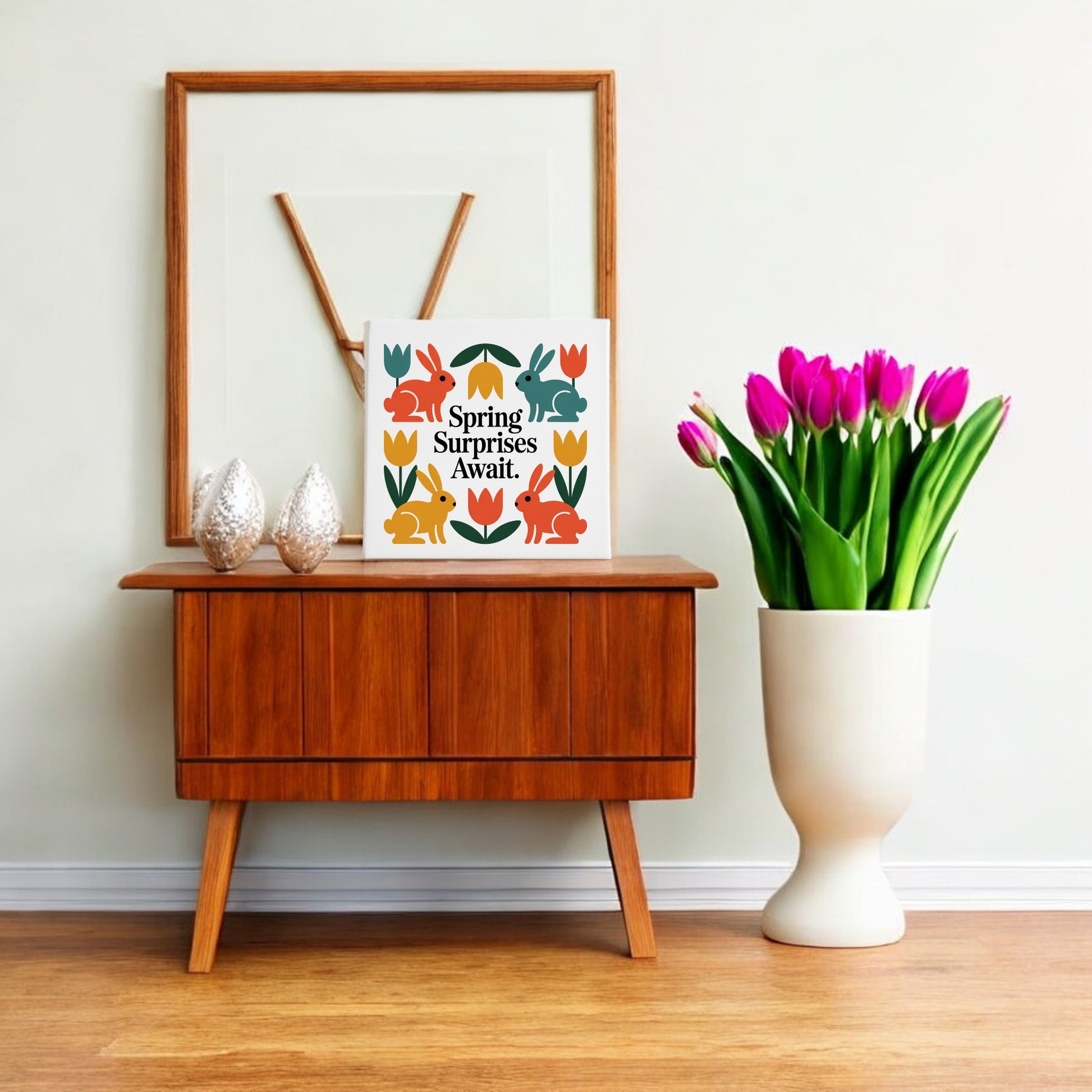 A wooden sideboard with decor sits by a wall. On top are two textured ornaments, Scandinavian-inspired art, and Mirabilia Boutique’s "Spring Surprises Await Canvas – 8x8 Colorful Bunnies." A white vase of pink tulips stands beside the sideboard.