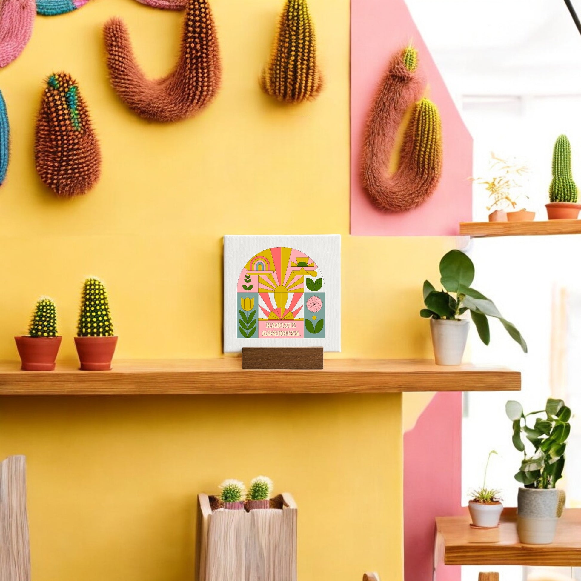 A modern, colorful room with yellow and pink walls showcases cacti, potted plants on a wooden shelf, textured hanging art, and the "Radiate Goodness Mini Canvas Sign with Base" by Mirabilia Boutique as cheerful encouragement decor.