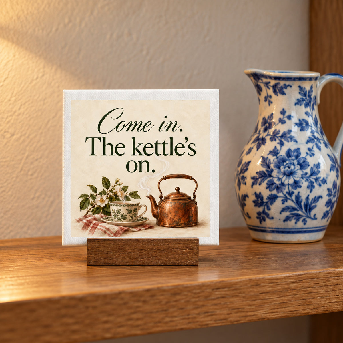 A Come In, The Kettle’s On Mini Canvas on a wooden stand reads, “Come in. The kettle’s on,” featuring an illustration of a teacup and a steaming copper kettle.