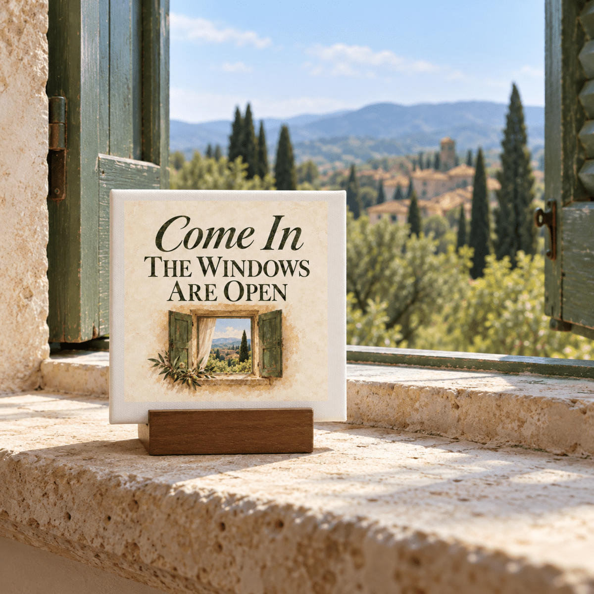 A Come In Mini Canvas Sign | Ochre & Cypress on a wooden stand sits on a stone windowsill, featuring an open window illustration, with countryside wall art of hills and trees in the background.
