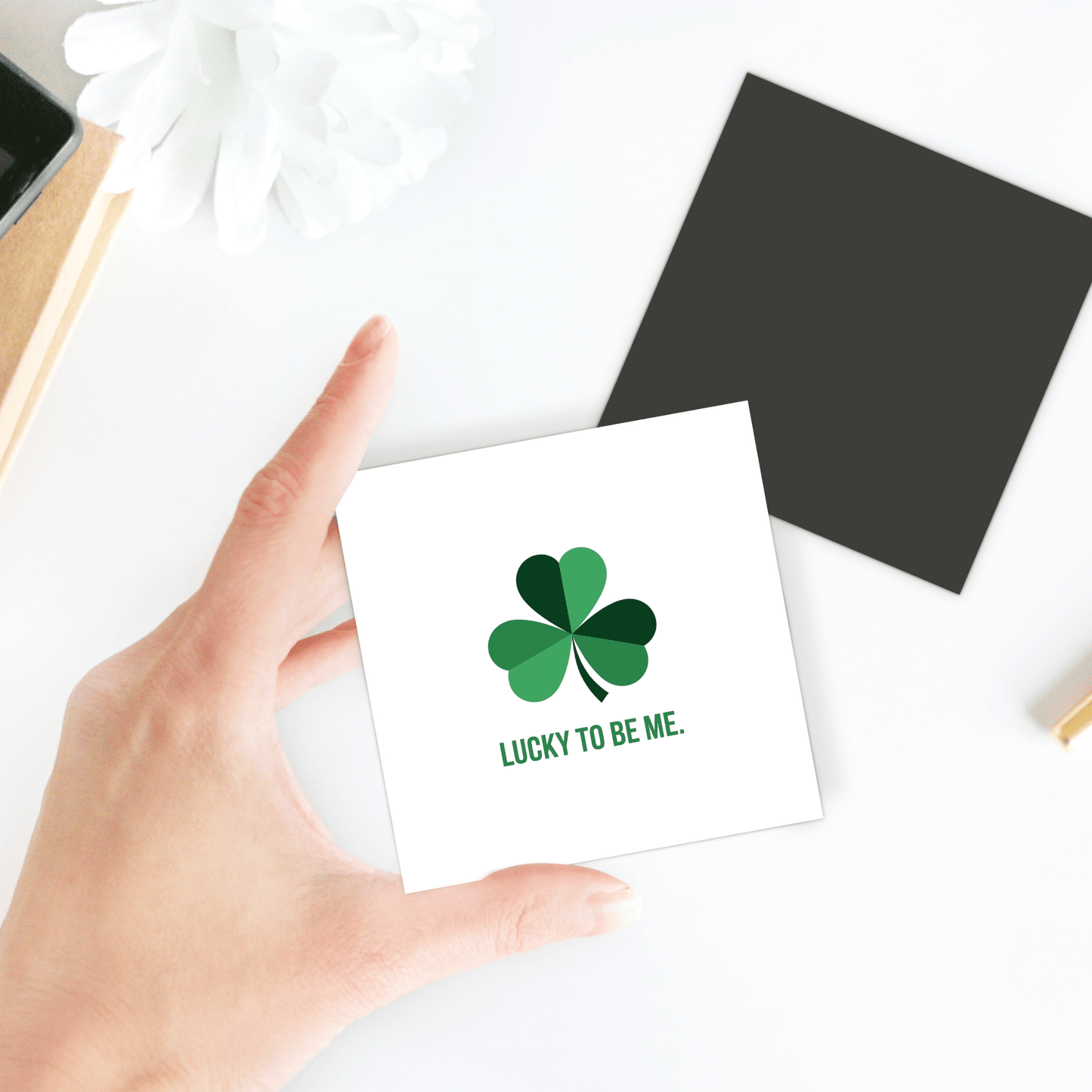 A hand holds the "Lucky to Be Me Shamrock Magnet" by Mirabilia Boutique, featuring a green four-leaf clover. Nearby, a black square and white flower add charm to this St. Patrick’s Day magnet display on a white surface.