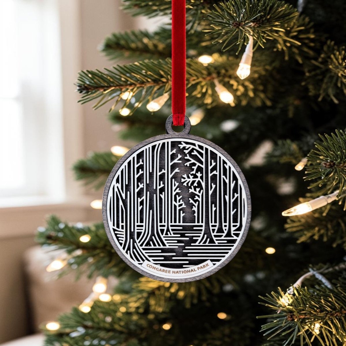 The Congaree National Park Christmas Ornament by Mirabilia Boutique—a handcrafted wooden piece featuring a detailed forest design and "Congaree National Park" inscribed at the bottom—hangs from a red ribbon on a Christmas tree adorned with white string lights, with a softly blurred window in the background, making it the perfect old-growth forest keepsake for your holiday décor.