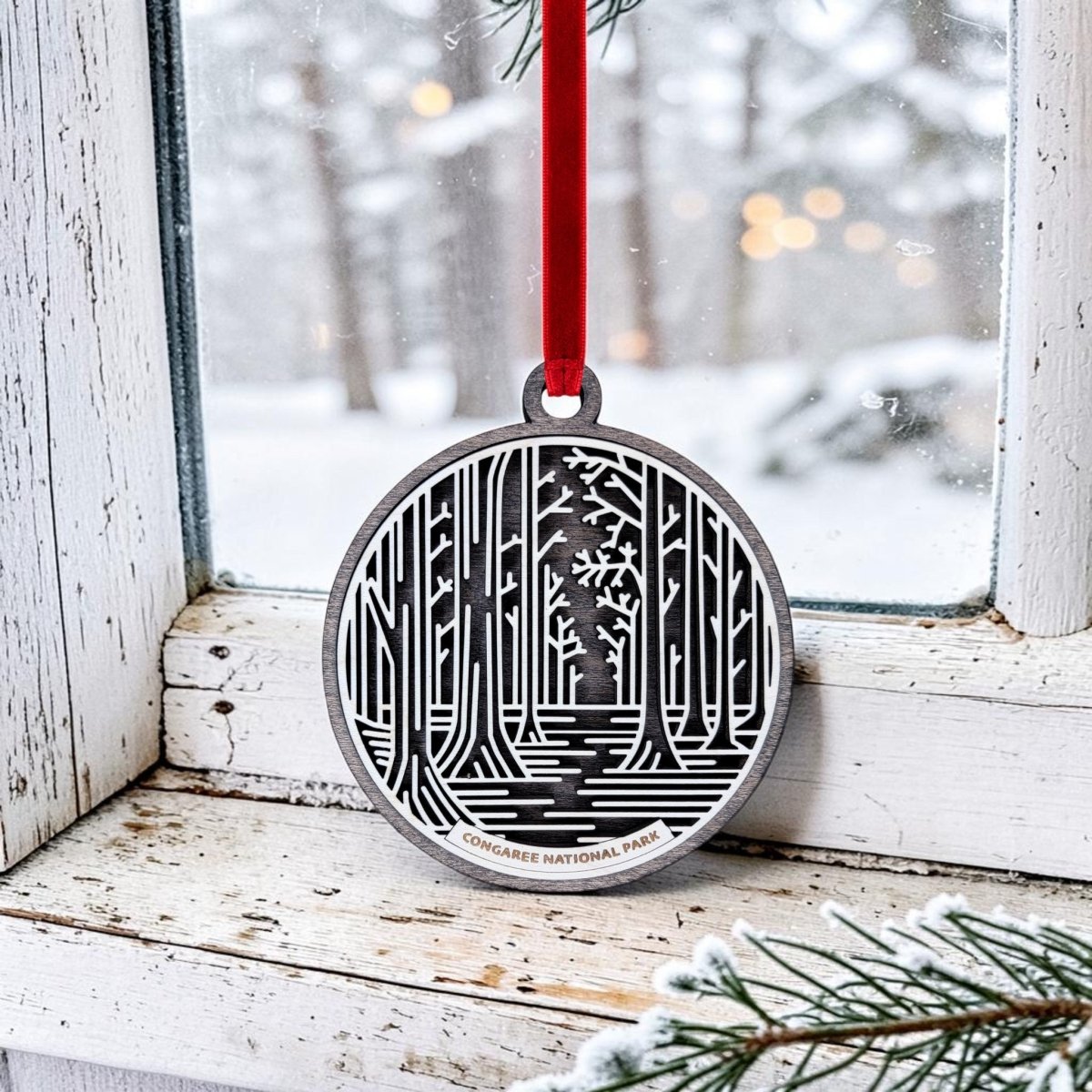 A Mirabilia Boutique Congaree National Park Christmas Ornament, featuring a handcrafted wooden forest design, hangs by a red ribbon on a snowy window ledge with snow-covered trees and blurred lights visible outside.