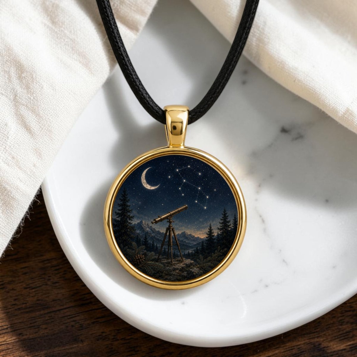 Introducing the Under the Constellations | Story Pendant Necklace by Mirabilia Boutique—a gold pendant on a black cord, adorned with intricate artwork of a telescope beneath a starry night sky featuring a crescent moon, constellation, trees, and mountains. This elegant piece is perfect for stargazing enthusiasts and jewelry lovers alike. Shown here resting on a white dish over a wooden surface.