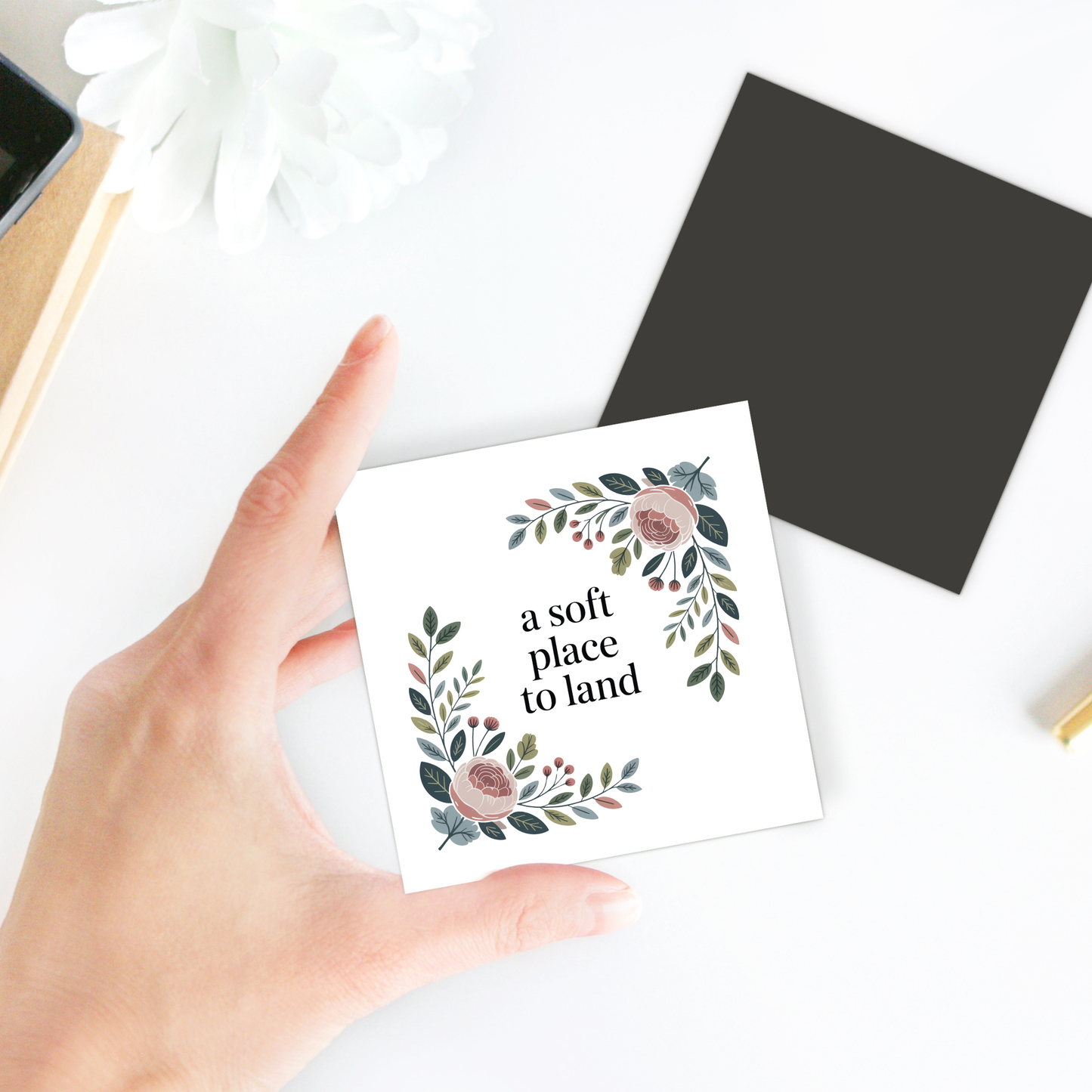 A hand holds the “A Soft Place to Land” floral magnet by Mirabilia Boutique, featuring botanical illustrations and a central quote. The charming display includes a black square, a flower, and hints of stationery in the background.