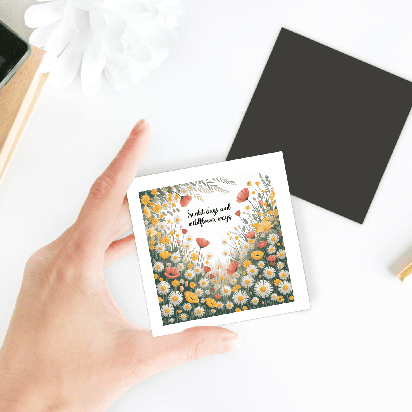 A hand holds the Sunlit Days and Wildflower Ways Magnet from Mirabilia Boutique, a vibrant floral summer fridge art card on a white surface near a blank black square and a white flower.