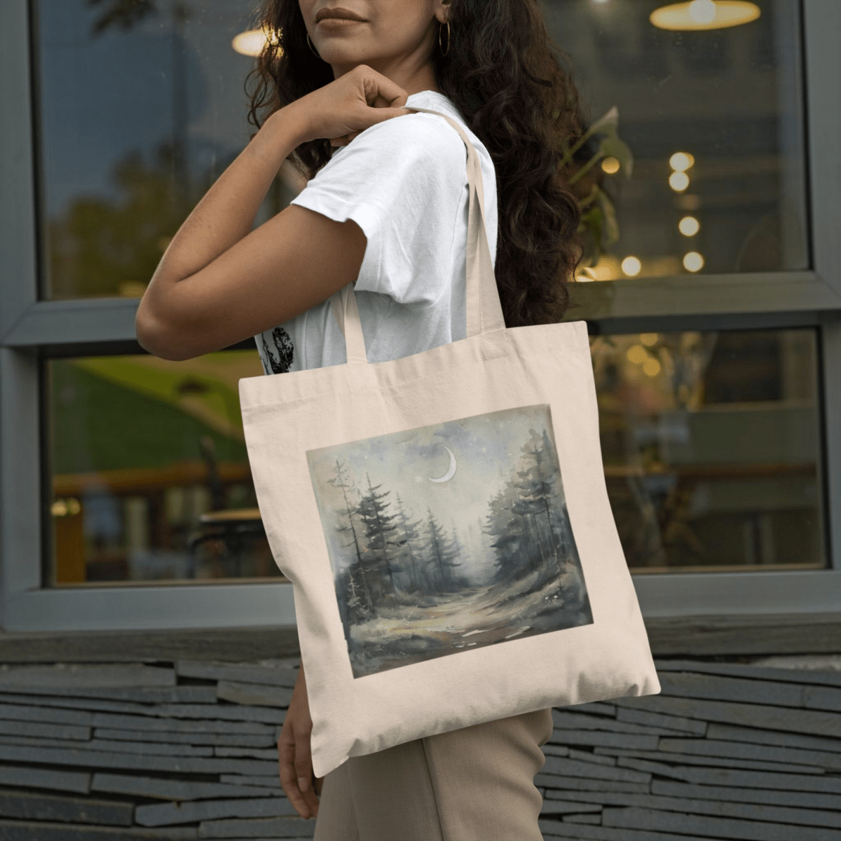 A woman stands outside by a window, holding the "Some Paths Are Meant to Be Wandered" tote bag from Mirabilia Boutique, which features a painted forest scene with pine trees, a crescent moon, and a misty path.