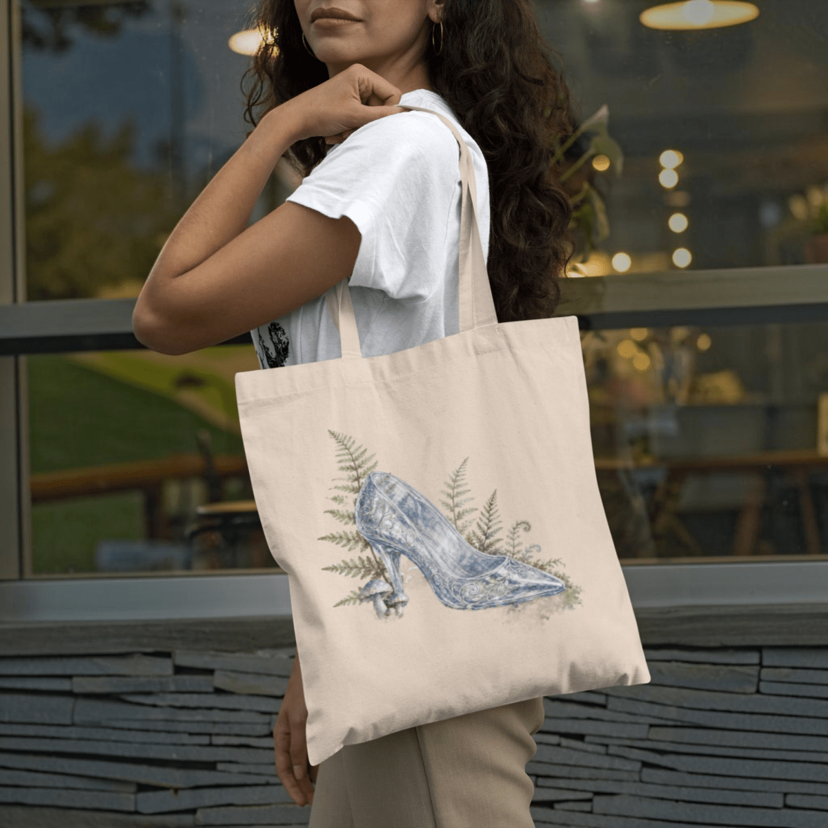 A woman stands outdoors near a glass window, holding the Enchanted Glass Slipper Canvas Tote Bag by Mirabilia Boutique—a beige tote adorned with an illustration of a glass slipper surrounded by ferns. This charming fairytale accessory is the perfect cottagecore piece.