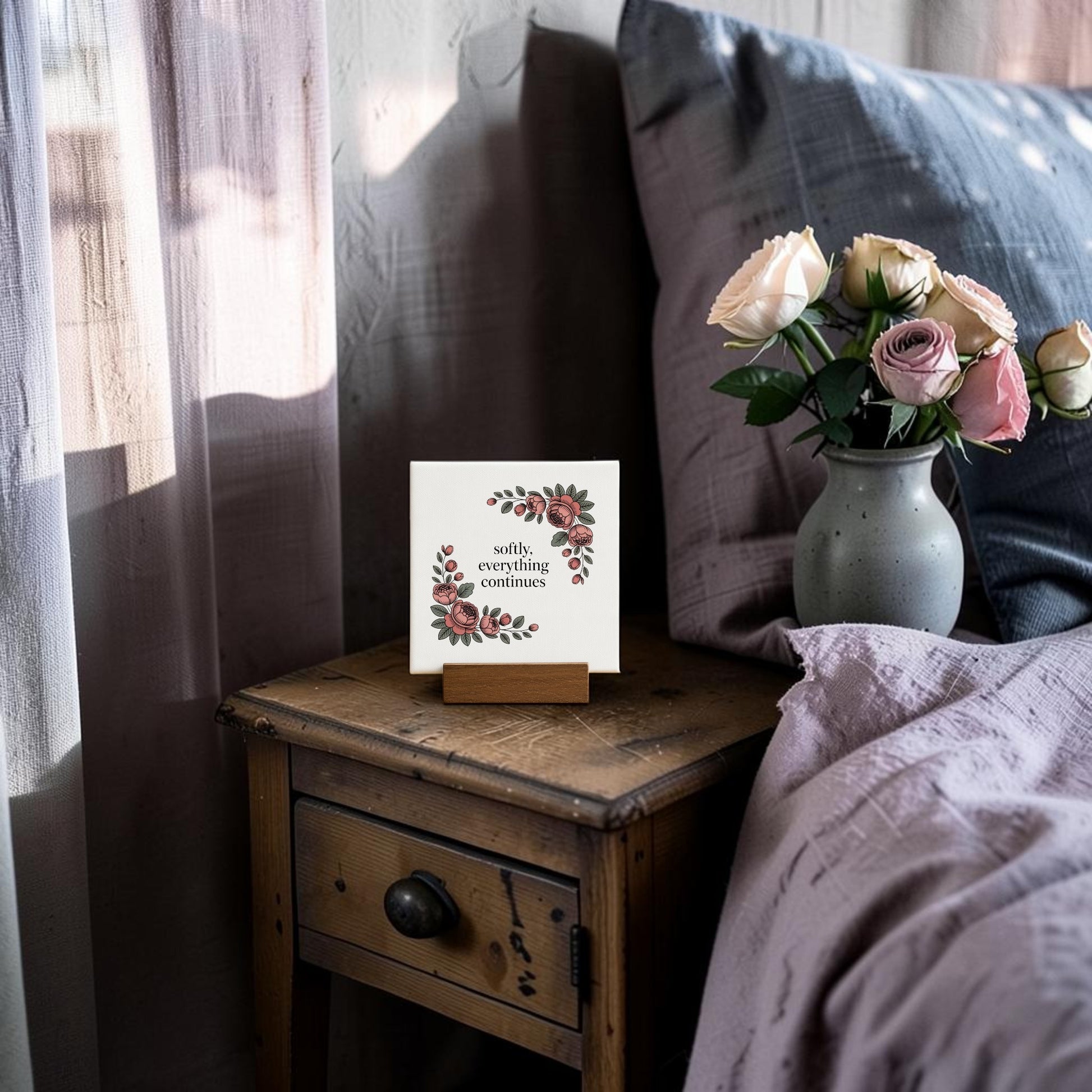 A small wooden bedside table holds a vase of pink and white roses and the Mirabilia Boutique "Softly, Everything Continues" Mini Sign with floral designs—an elegant, meaningful gift—beside a bed with a mauve blanket.