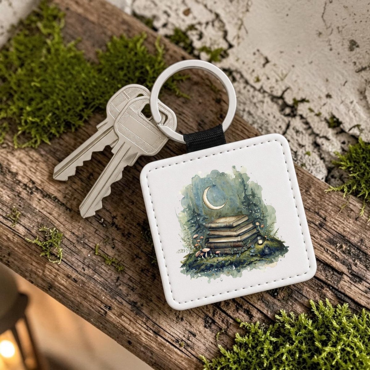 Add a touch of cottagecore charm to your everyday essentials with the Bookish Woodland Keychain by Mirabilia Boutique—a set of two silver keys attached to a square fob adorned with enchanting artwork of stacked books, a crescent moon, and forest plants. Perfect for book lovers, this magical accessory is pictured resting on a wooden surface surrounded by lush green moss.