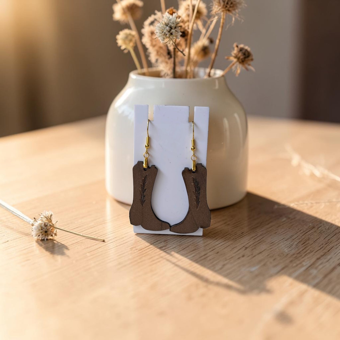 The Cowboy Boot Earrings by Mirabilia Boutique, crafted from lasercut walnut wood, are displayed on a white card in front of a cream vase with dried flowers on a rustic table washed in warm natural light—perfect for western style jewelry lovers.