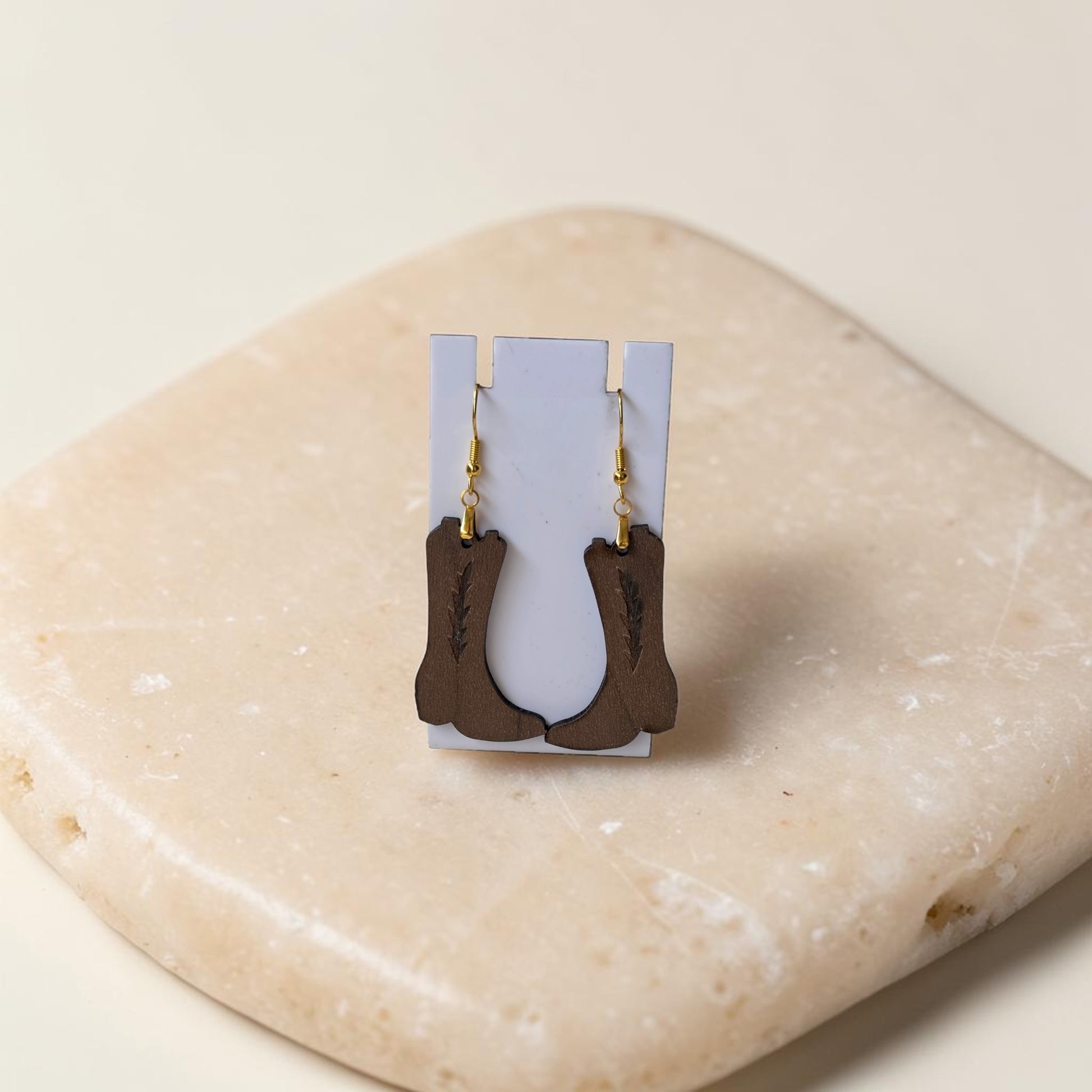 Add western flair to any look with Mirabilia Boutique’s Cowboy Boot Earrings, lasercut from walnut wood and finished with gold hooks, displayed on a white card atop a beige stone for a rustic touch.