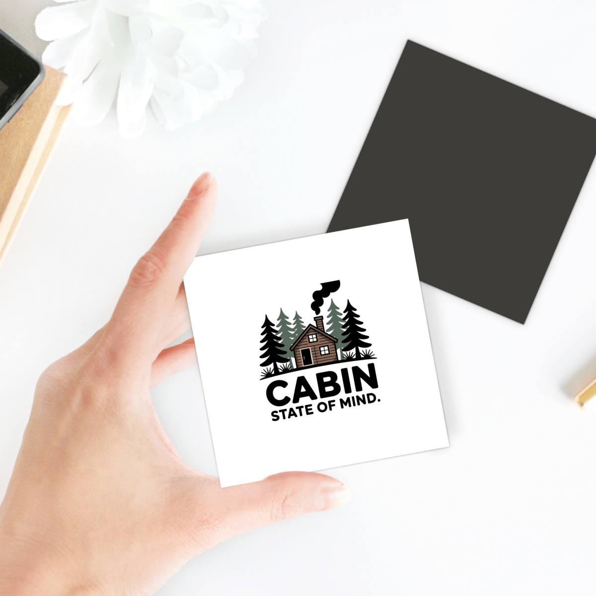 A hand holds the "Cabin State of Mind Magnet" by Mirabilia Boutique, featuring a cabin among trees with chimney smoke. This rustic cabin decor piece is a perfect home accessory, shown with desk items and a black square in the background.