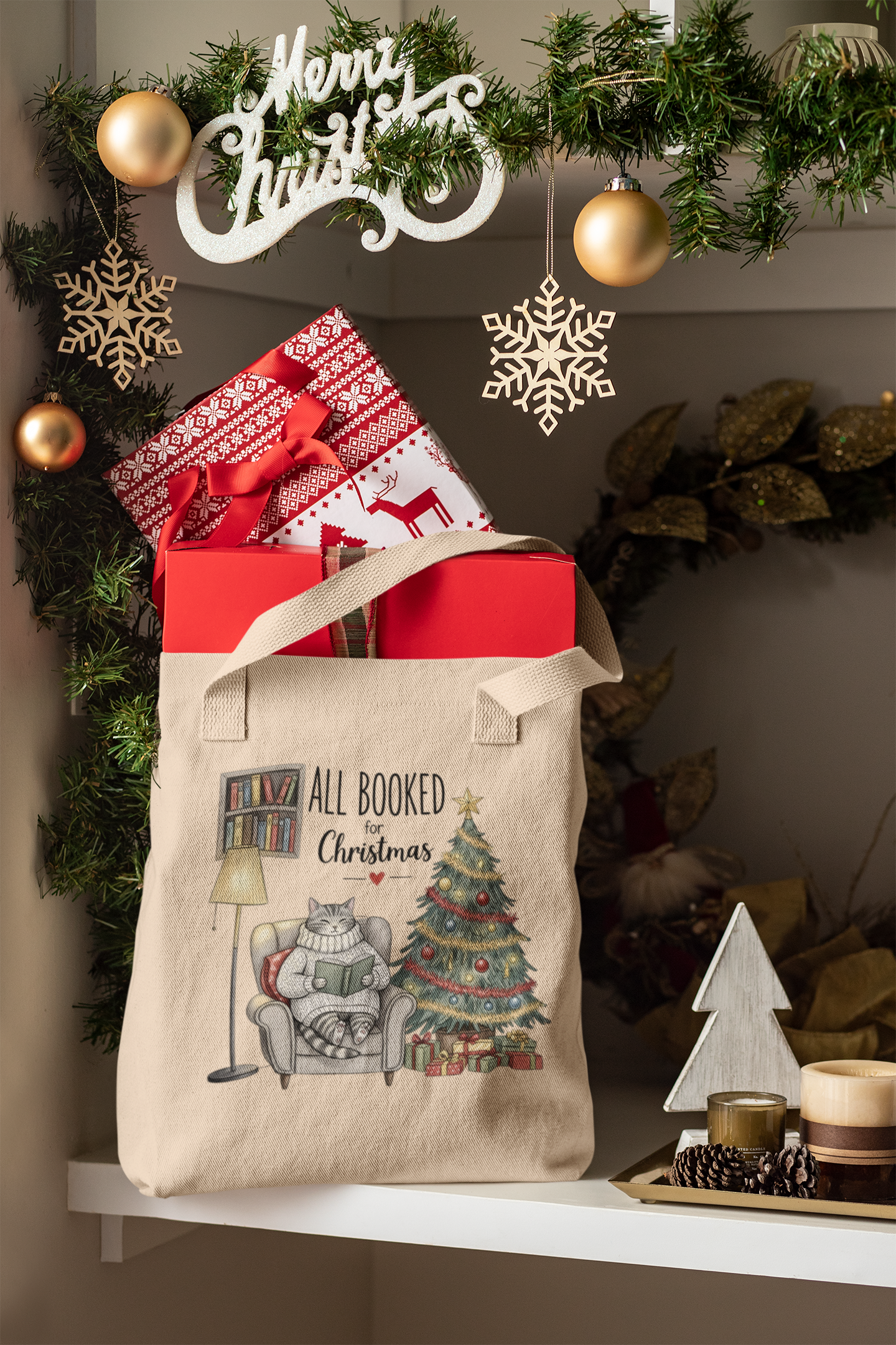 A festive "All Booked for Christmas Cat Tote Bag" by Mirabilia Boutique sits on a shelf with wrapped gifts, golden ornaments, greenery, and wooden holiday decor. The tote features a cat, books, Christmas tree, and presents.