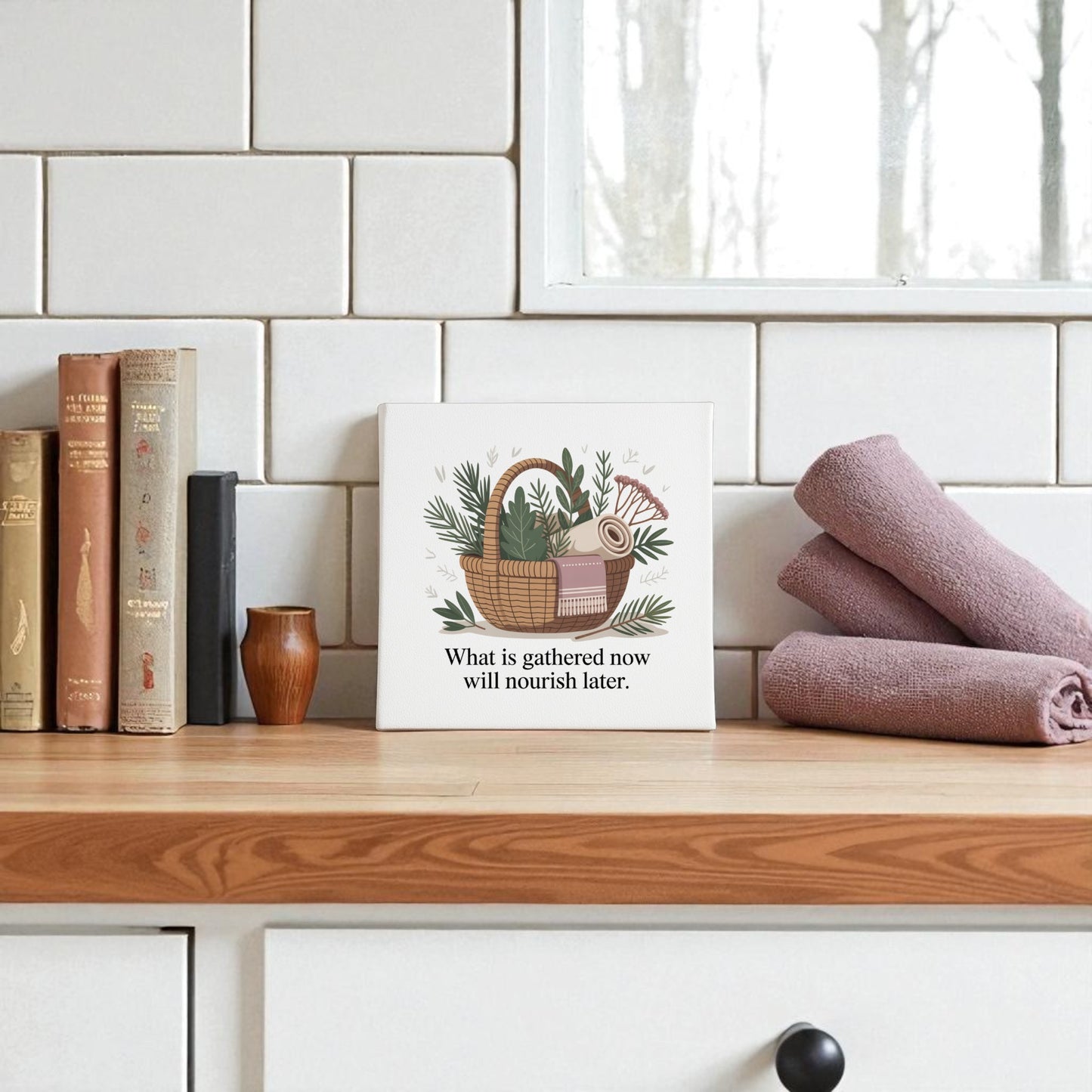 A kitchen counter styled with books, a vase, purple towels, and the Mirabilia Boutique Herbal Basket Canvas Art featuring “What Is Gathered Quote Print” brings a slow living vibe to white tiles.