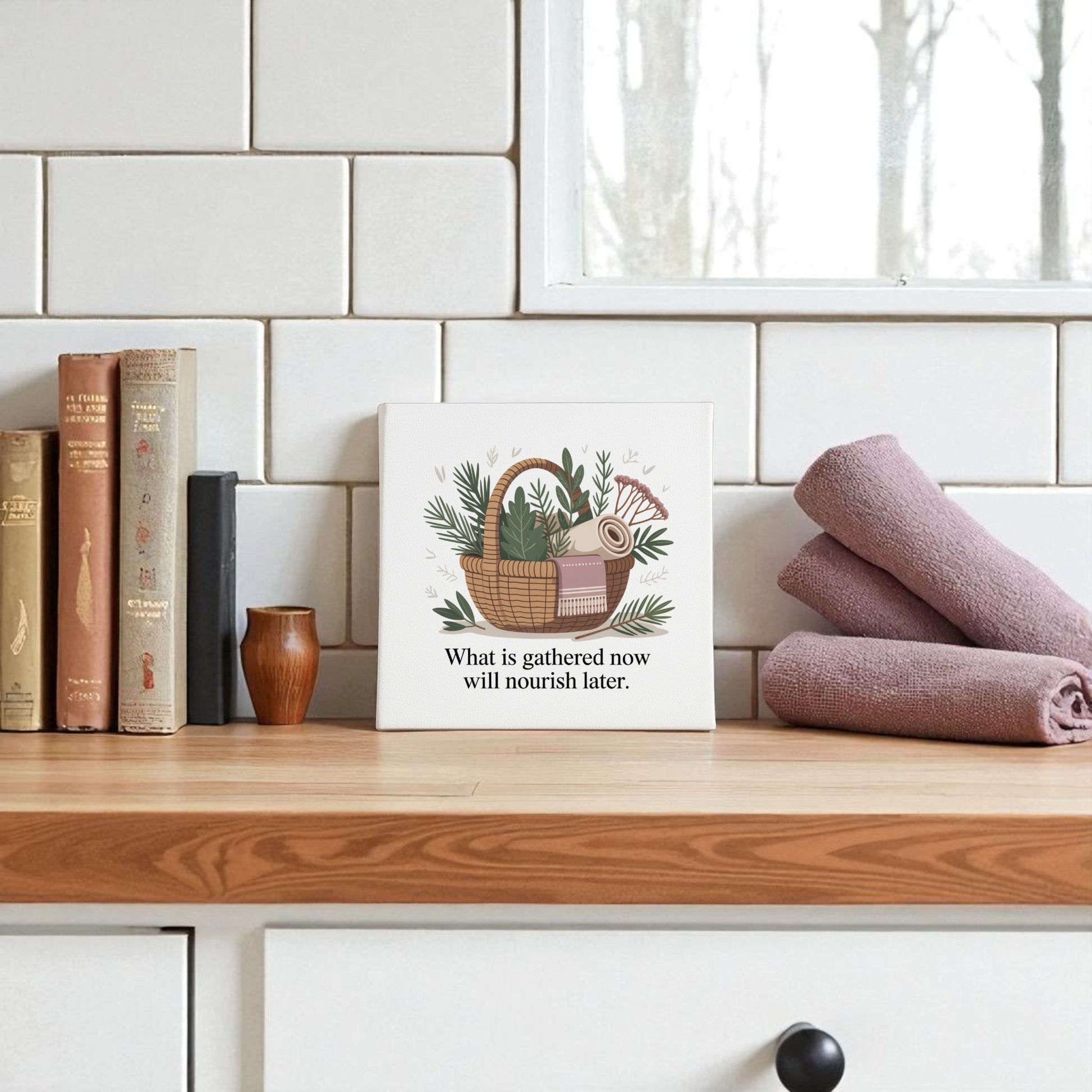 A kitchen counter styled with books, a vase, purple towels, and the Mirabilia Boutique Herbal Basket Canvas Art featuring “What Is Gathered Quote Print” brings a slow living vibe to white tiles.