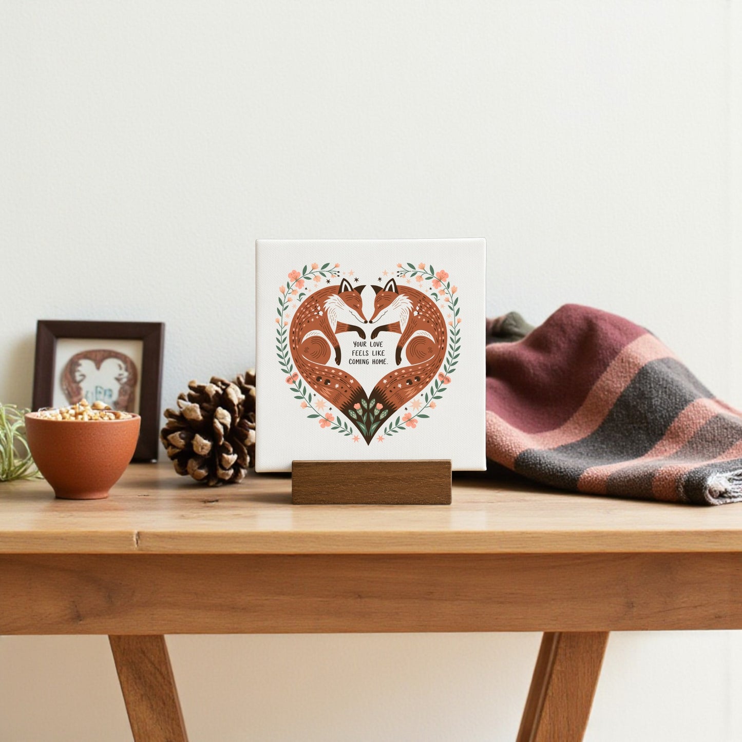 A wooden table displays a Mirabilia Boutique Fox Love Mini Canvas with the phrase “Your Love Feels Like Home,” a pinecone, a potted plant, and a folded plaid blanket as part of a cozy, woodland-inspired decor setup.