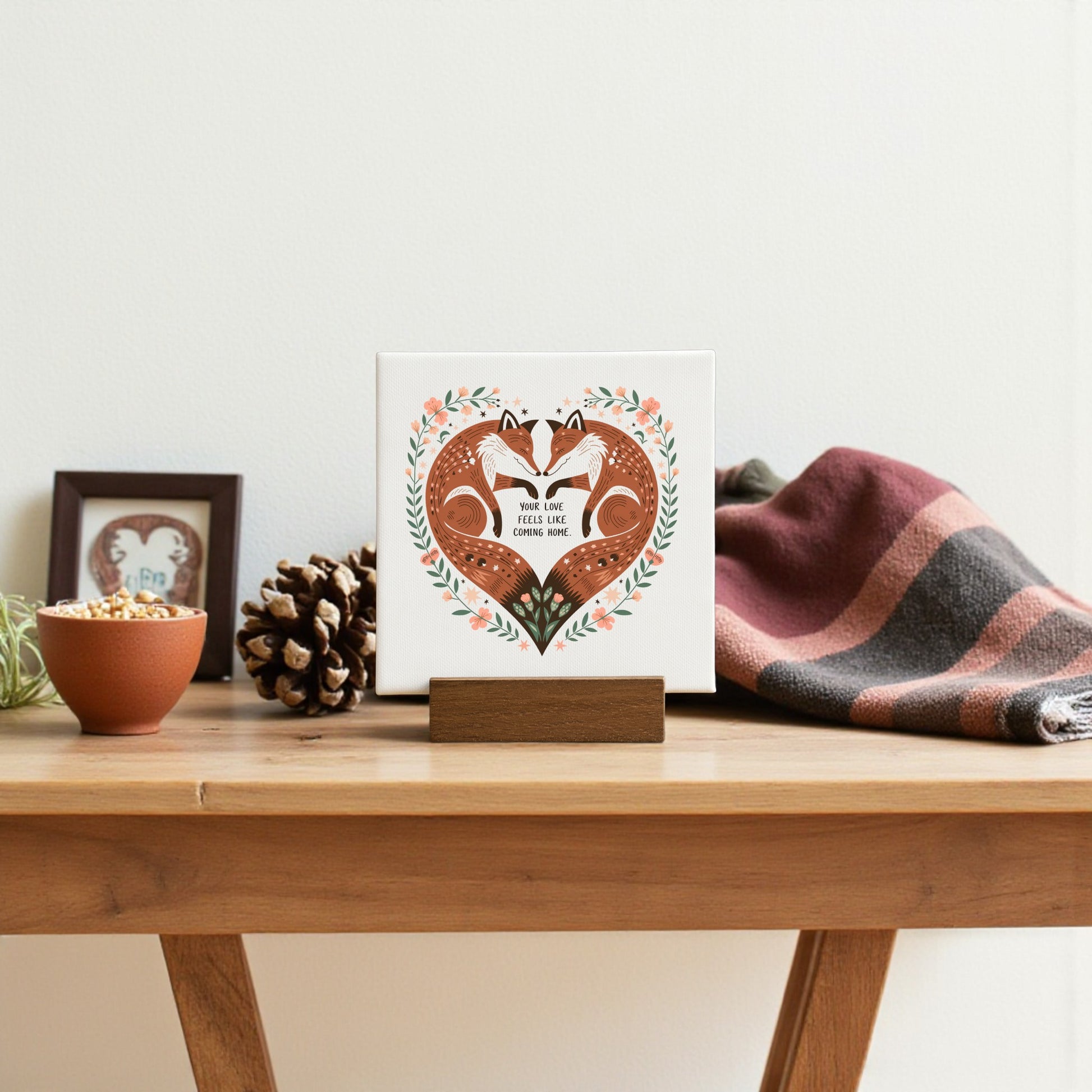 A wooden table displays a Mirabilia Boutique Fox Love Mini Canvas with the phrase “Your Love Feels Like Home,” a pinecone, a potted plant, and a folded plaid blanket as part of a cozy, woodland-inspired decor setup.